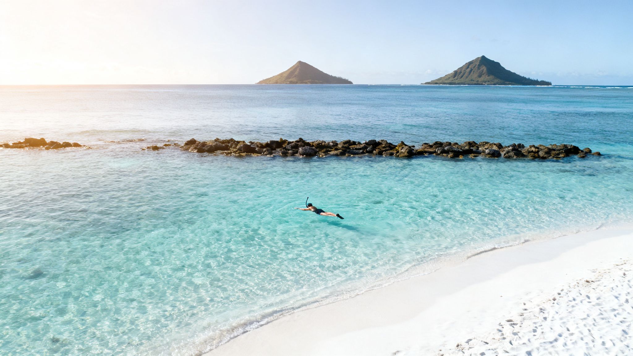 Aerial view of a person snorkeling in clear turquoise water near a white sandy beach with islands.