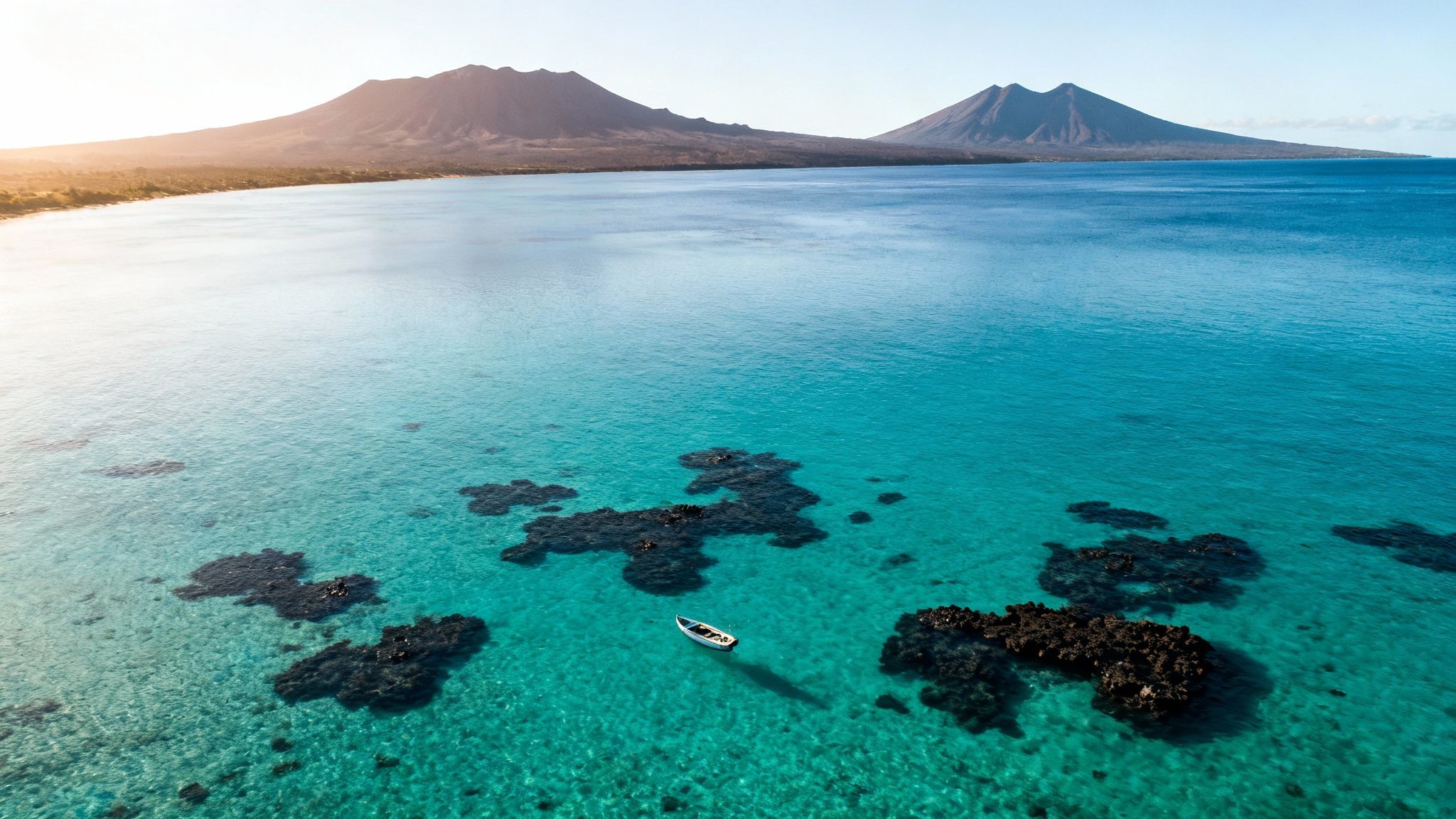 Aerial view of a white boat over vibrant turquoise water with coral reefs and mountains.