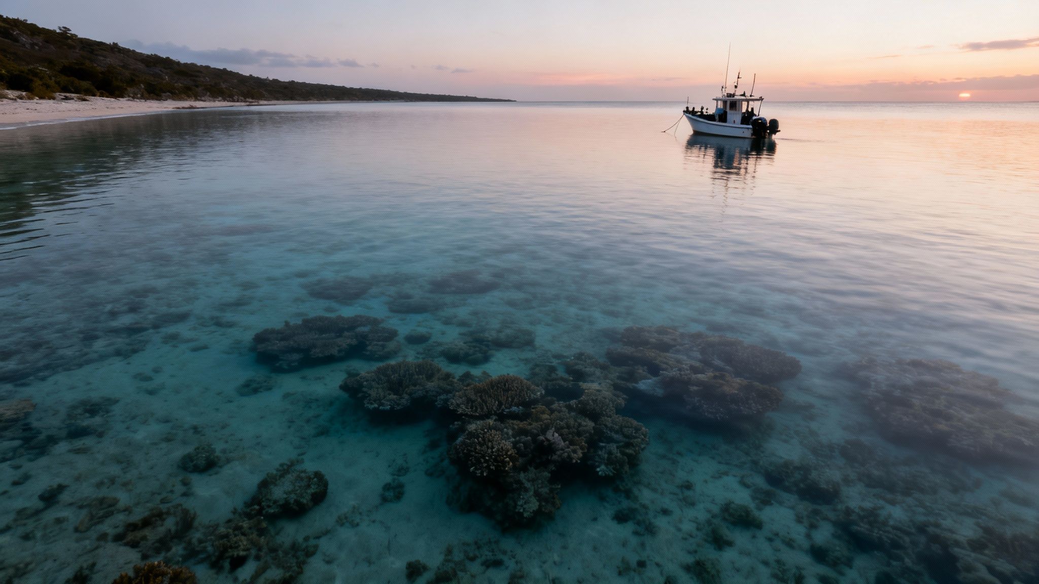 A tranquil sunset over a clear turquoise ocean with a fishing boat, sandy beach, and visible coral reefs.