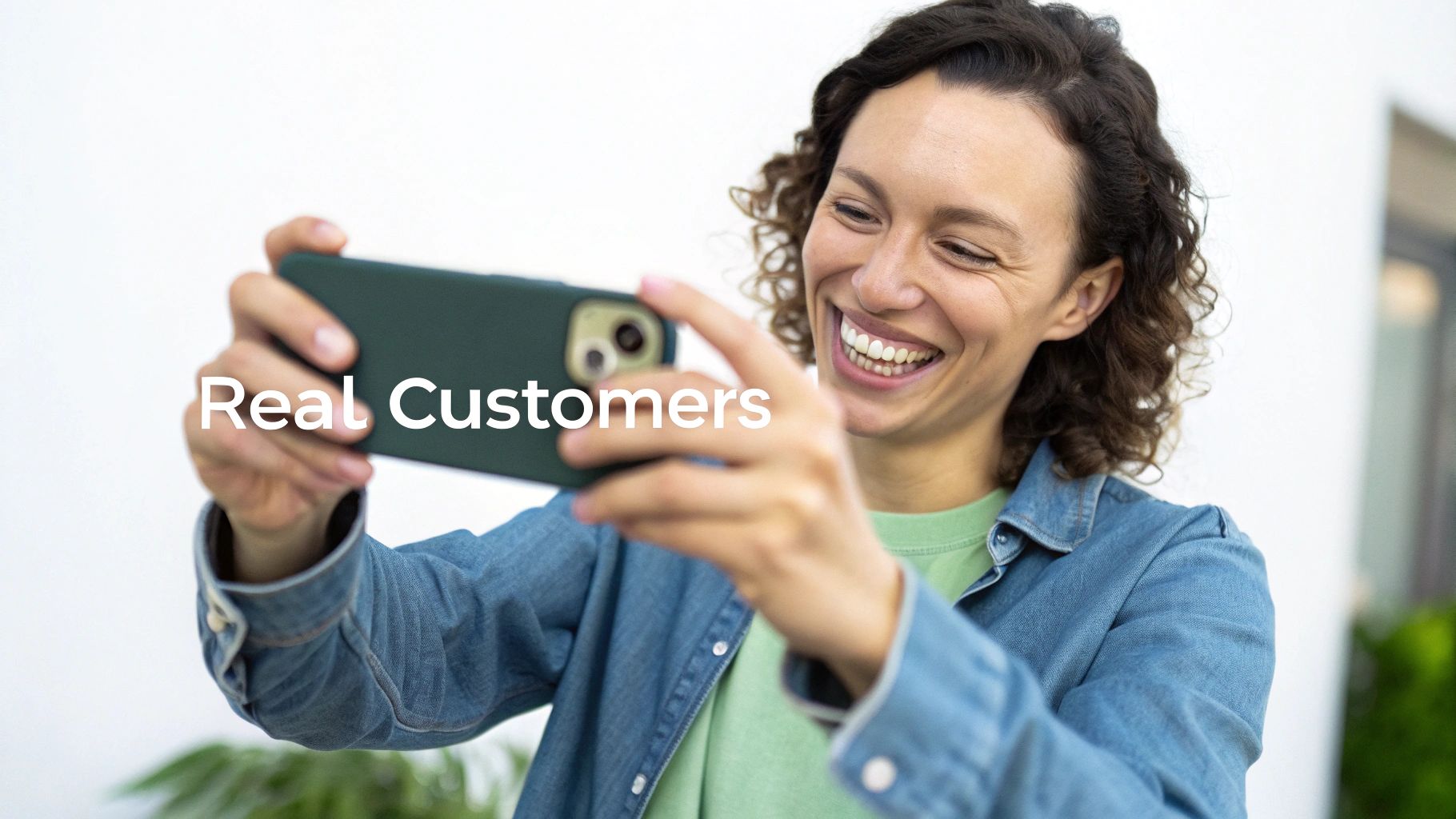 A happy woman with curly hair smiles brightly while holding a smartphone horizontally to take a photo, with 'Real Customers' text.