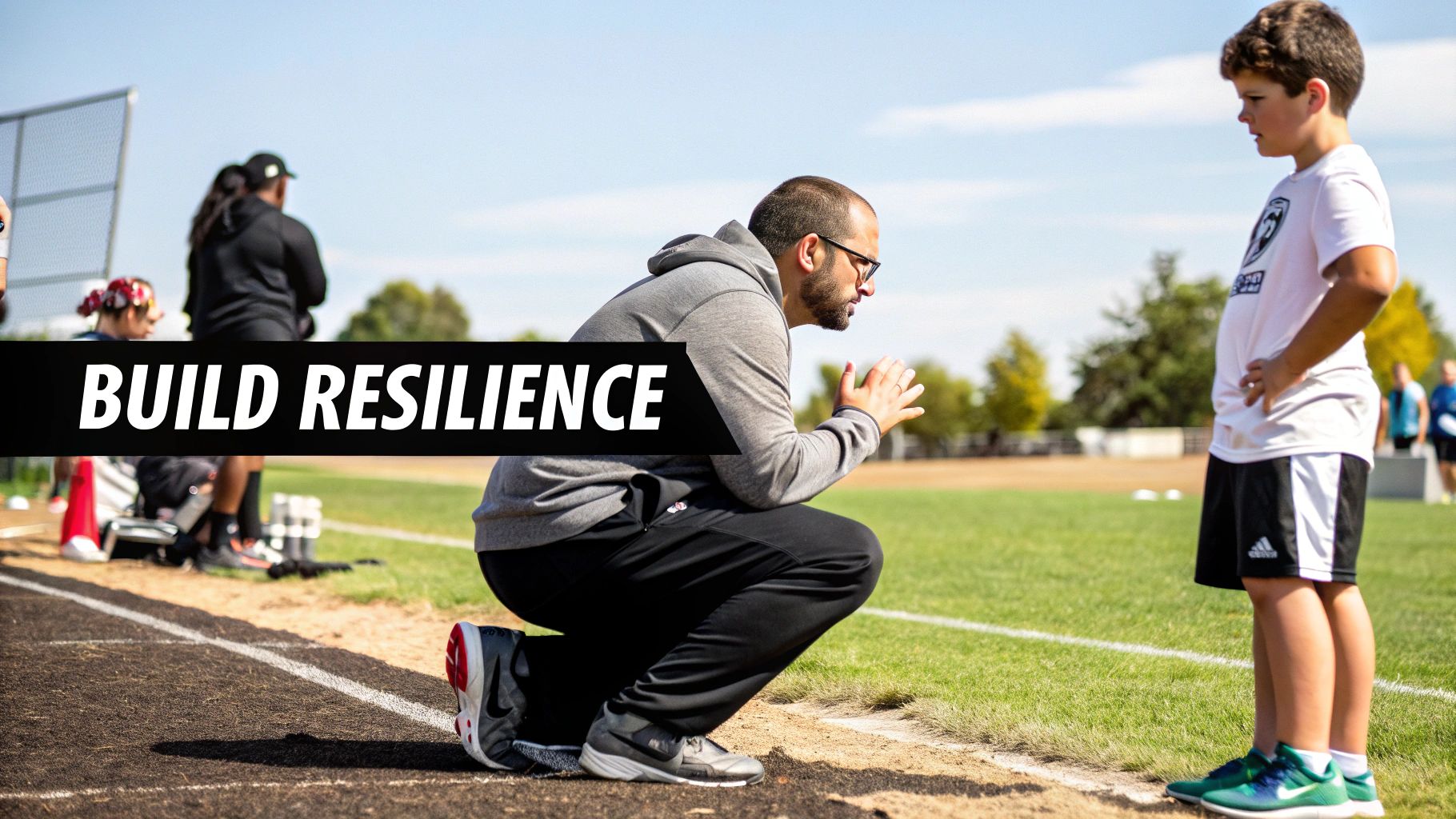 A male coach kneels on a track, intently talking to a young male athlete on a sunny sports field.