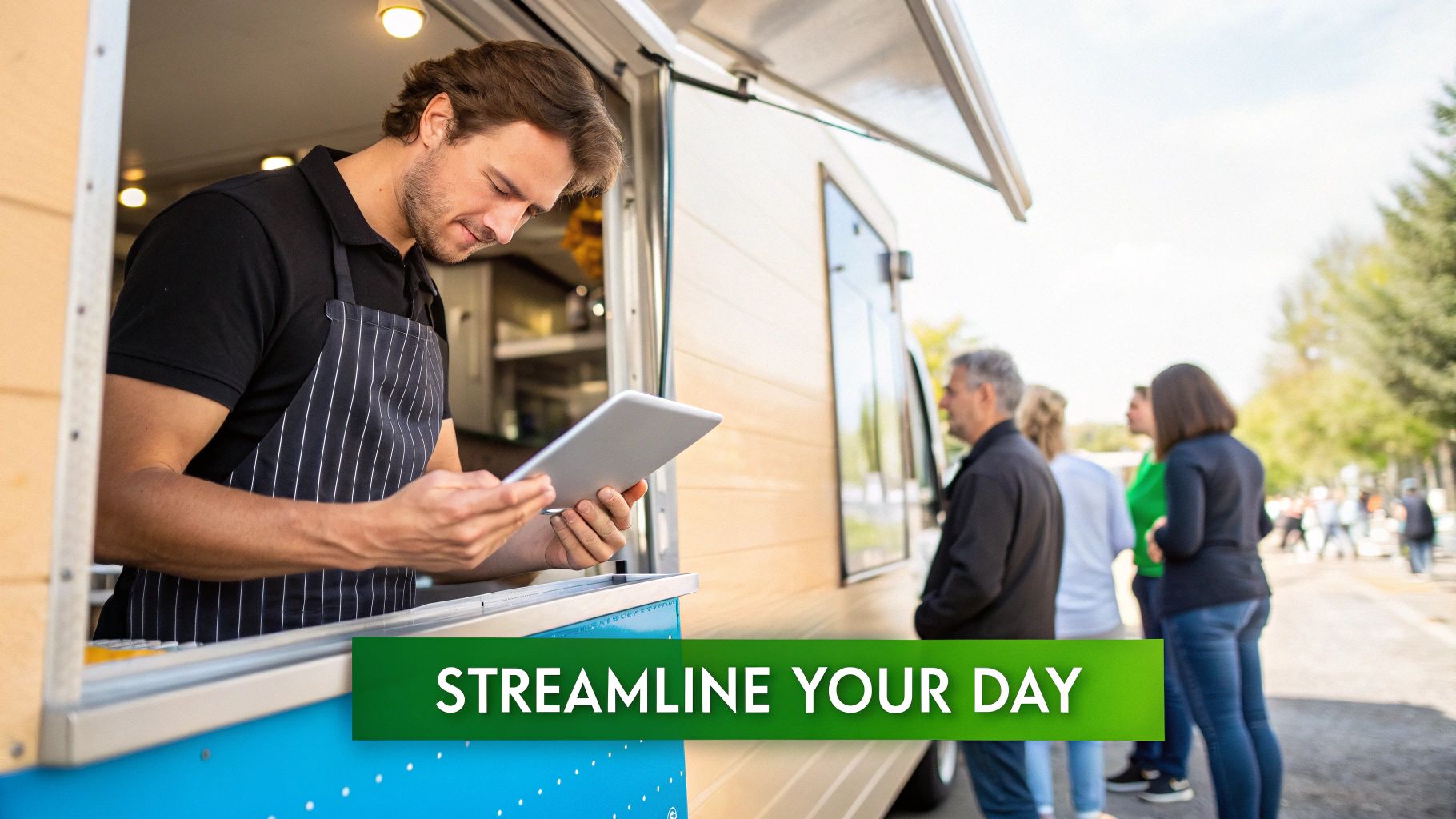 A male food truck vendor in an apron uses a tablet while customers wait in line.