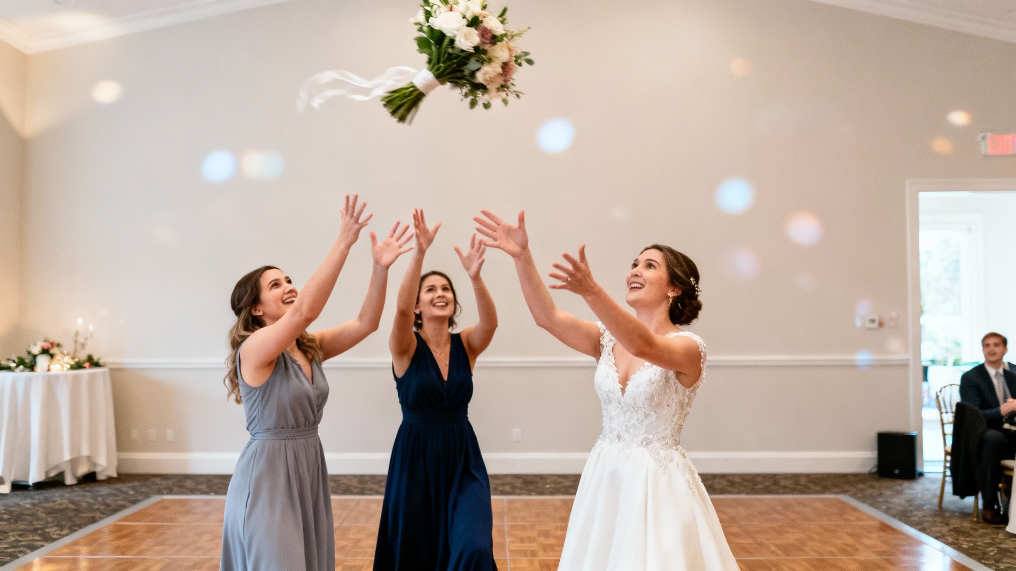 Three smiling women, including a bride, reaching for a tossed wedding bouquet indoors.