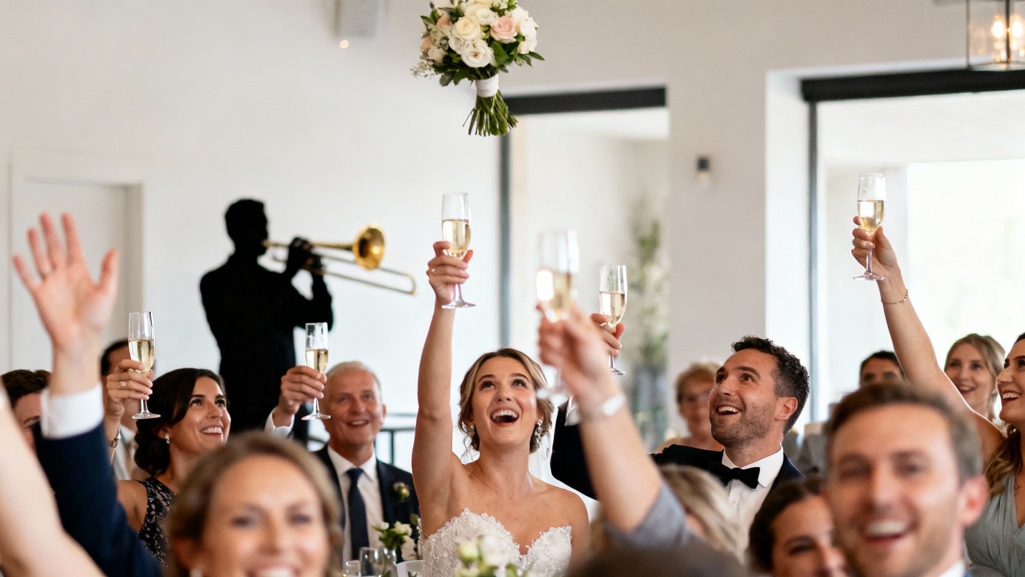Joyful bride tosses her wedding bouquet while excited guests raise champagne glasses.