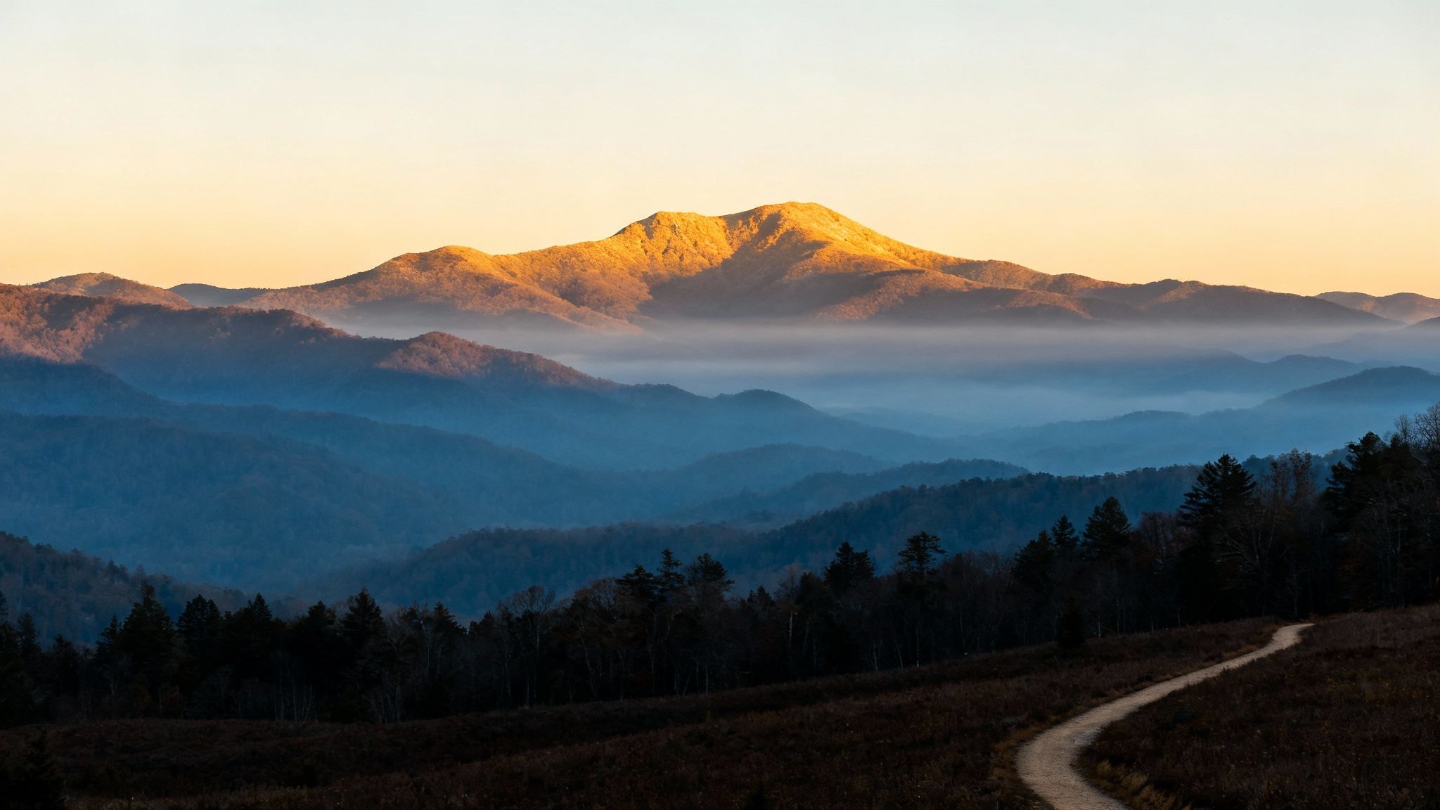 Golden light illuminates a majestic mountain peak above misty blue layers and a winding path.