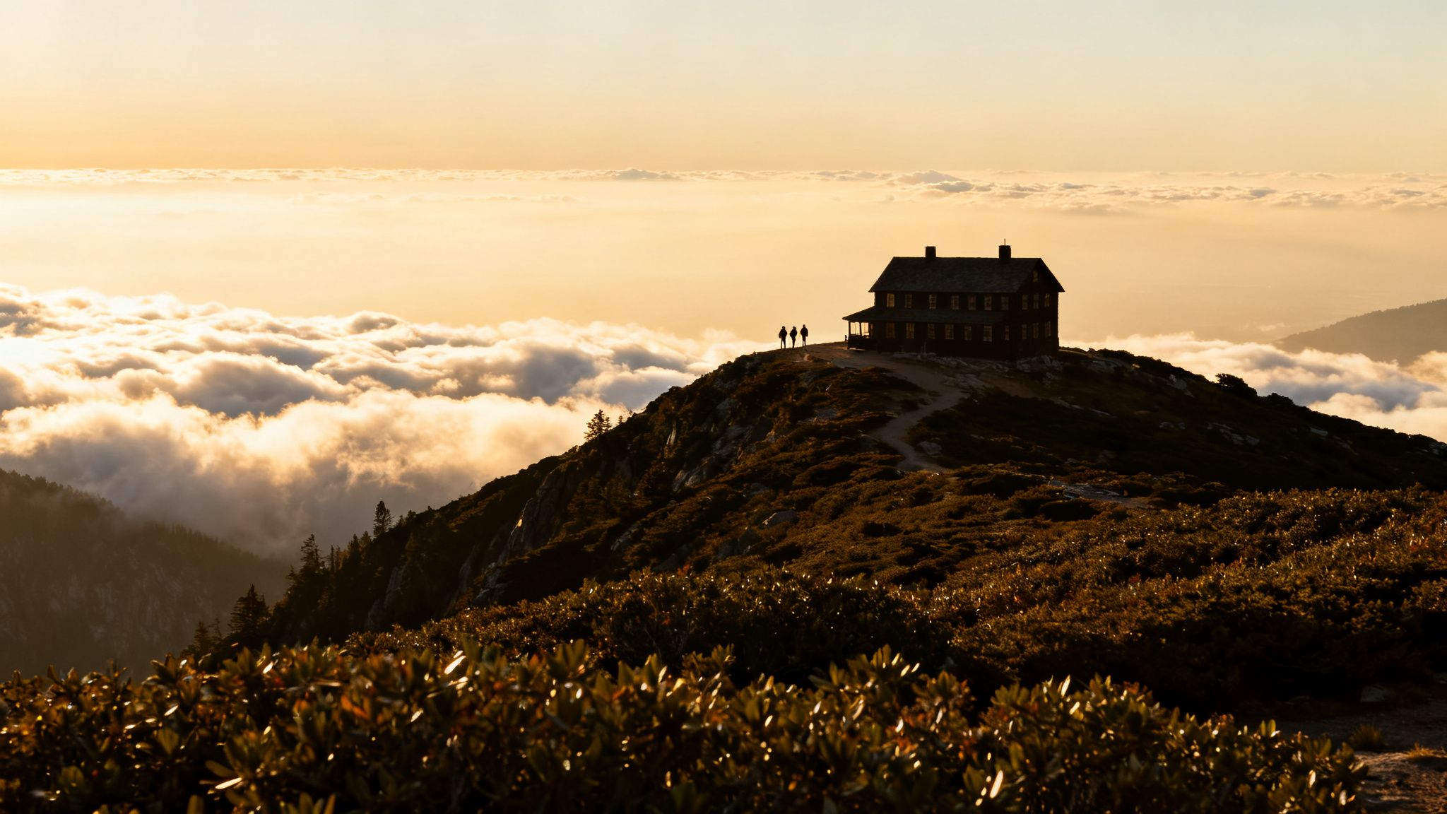 Golden hour view of a mountain hut and hikers above clouds on a scenic mountain.
