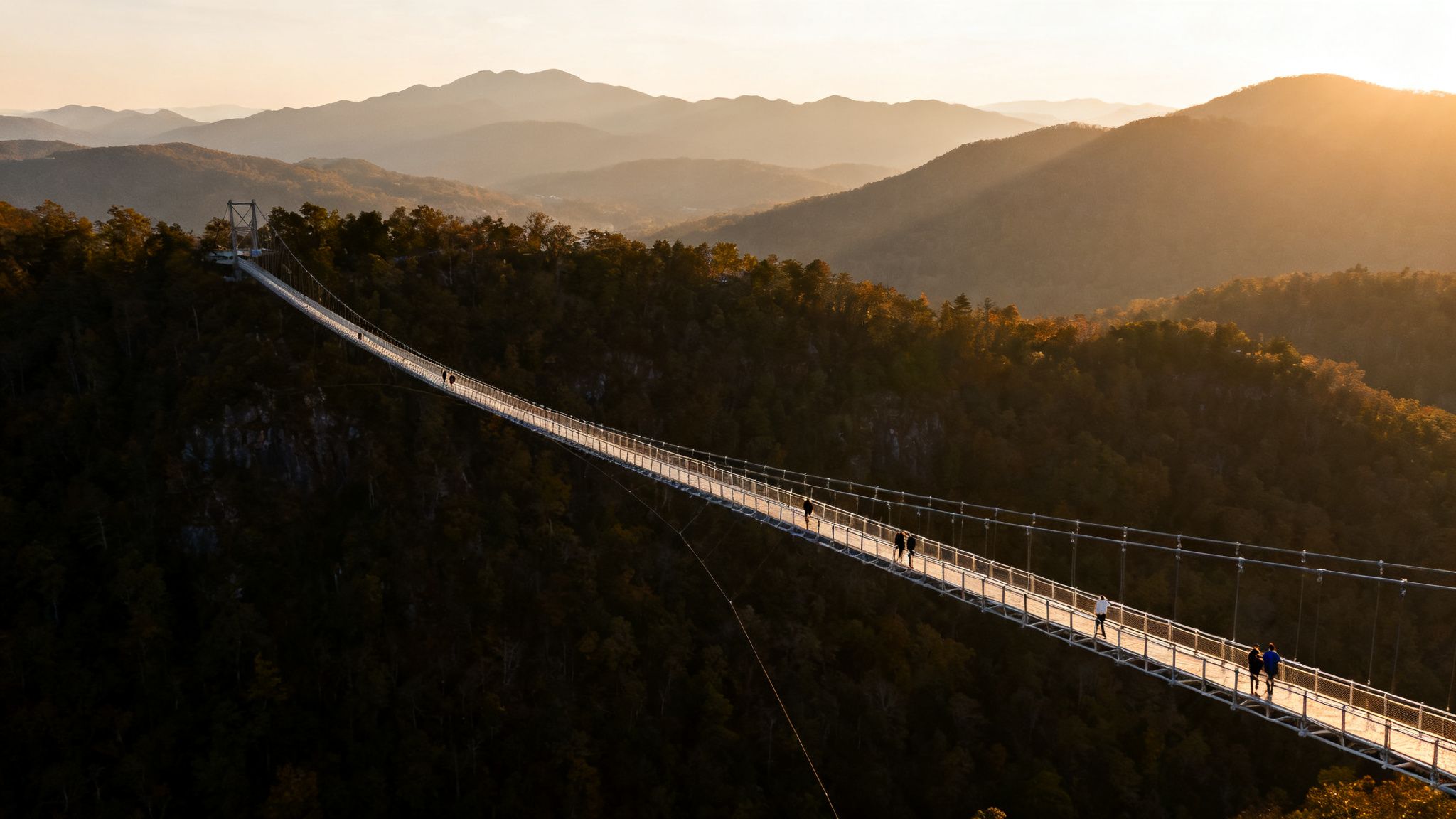 A long suspension bridge illuminated by golden sunset light over a forested valley with distant mountains.