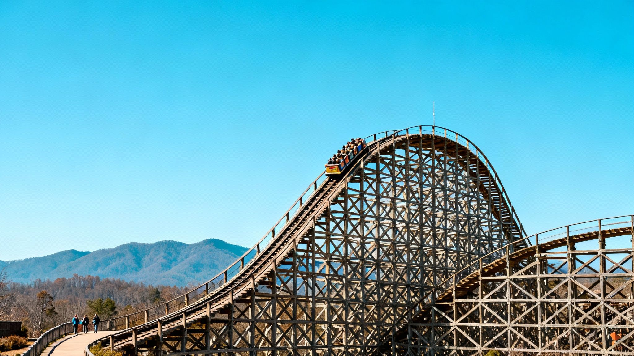 A wooden roller coaster with a full train ascends a steep hill under a clear blue sky.