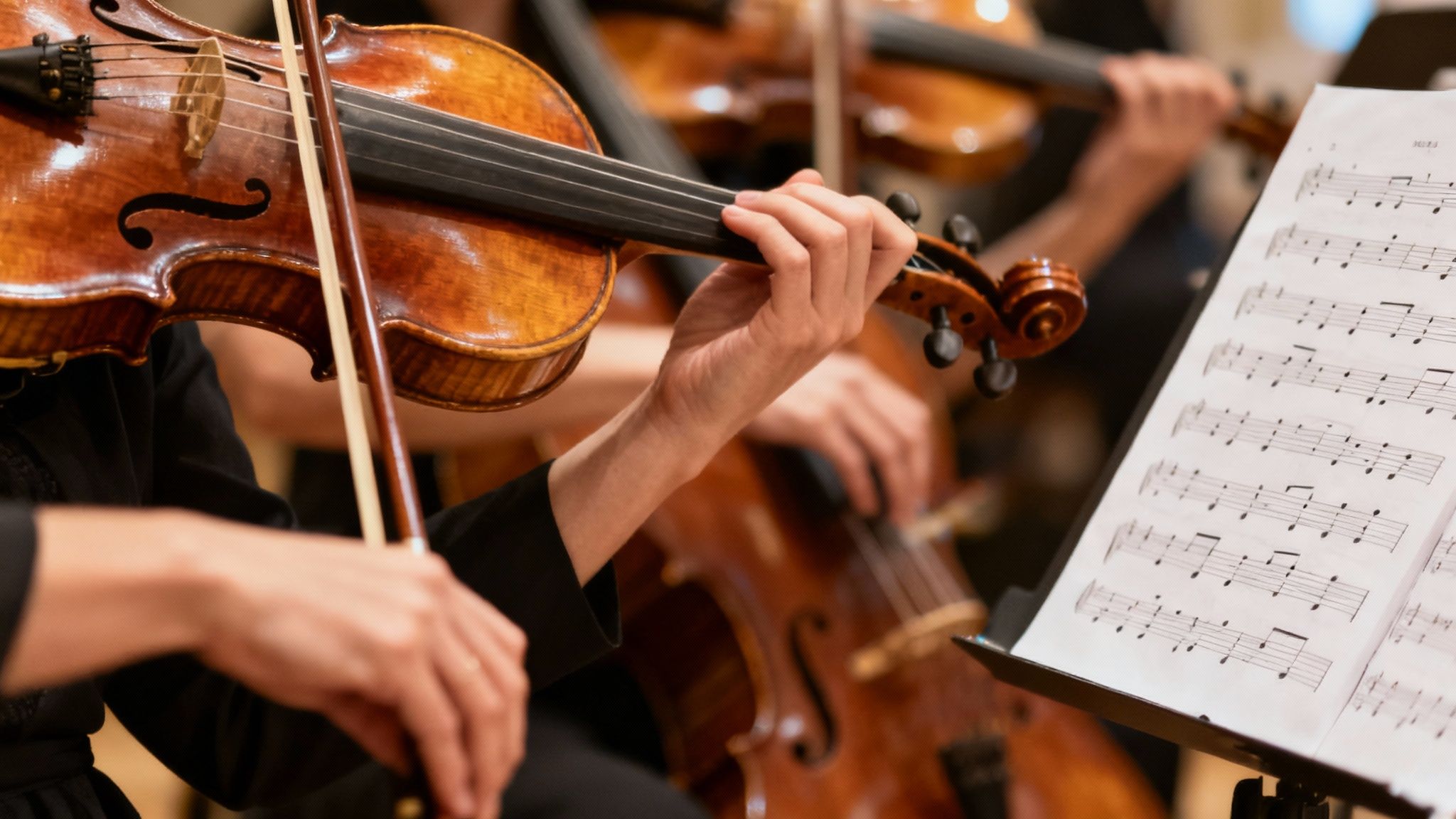 Close-up of musicians playing violins and a cello in an orchestra, with sheet music visible.