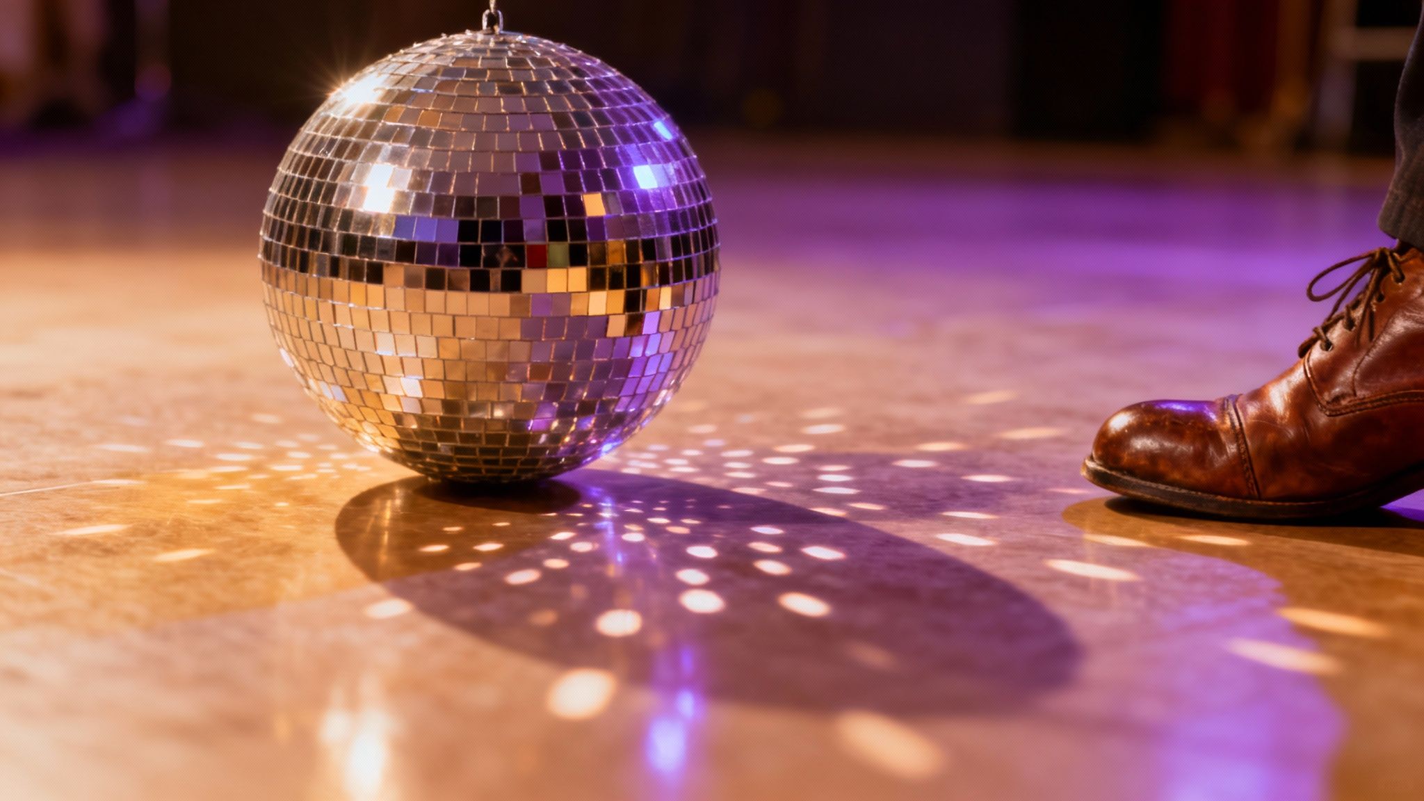 A shimmering disco ball reflects colorful lights on a dance floor next to a person's brown shoe.