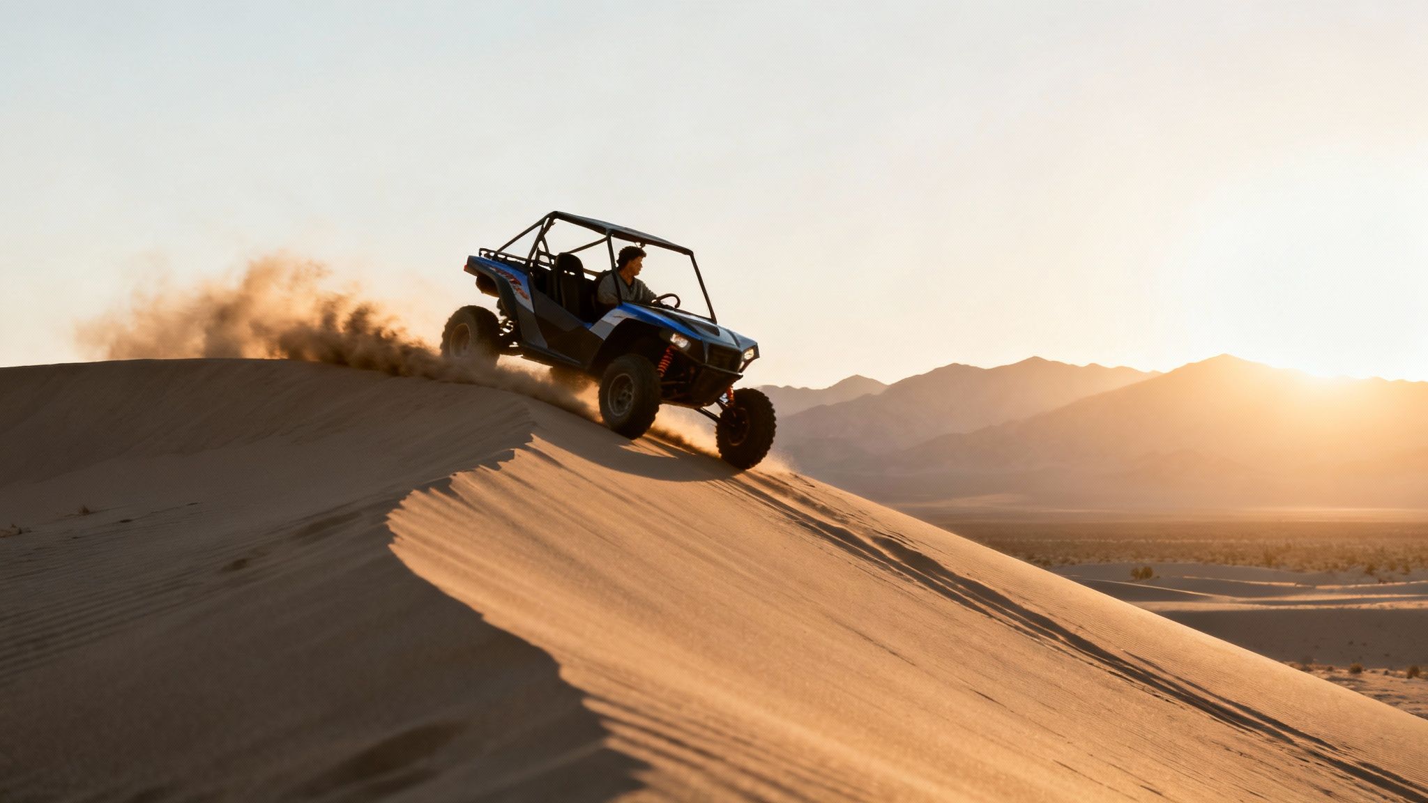 Person driving a blue UTV down a sand dune in the desert during golden hour.