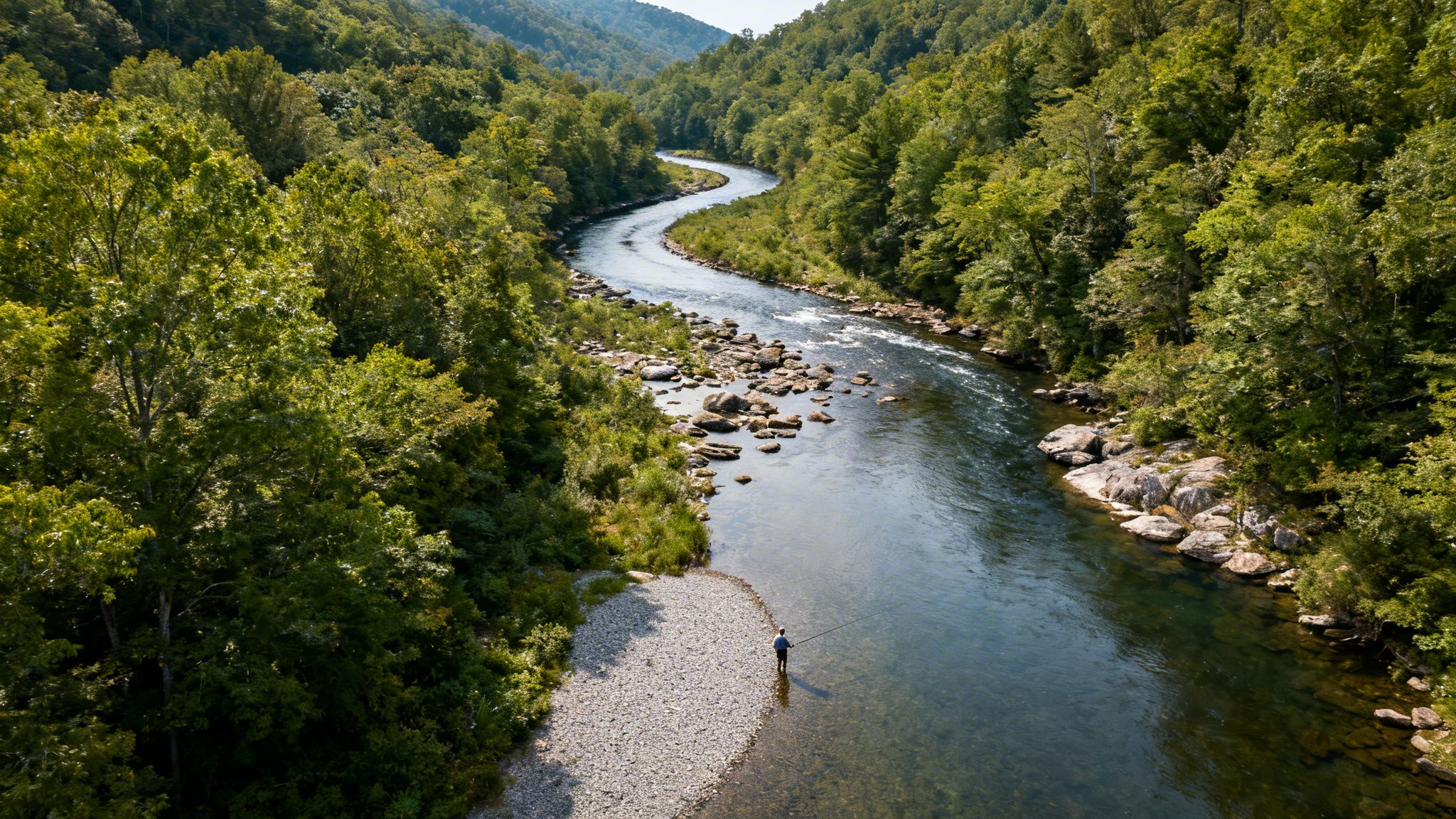 An aerial view shows a winding river through a lush green forest, with a person fly fishing.