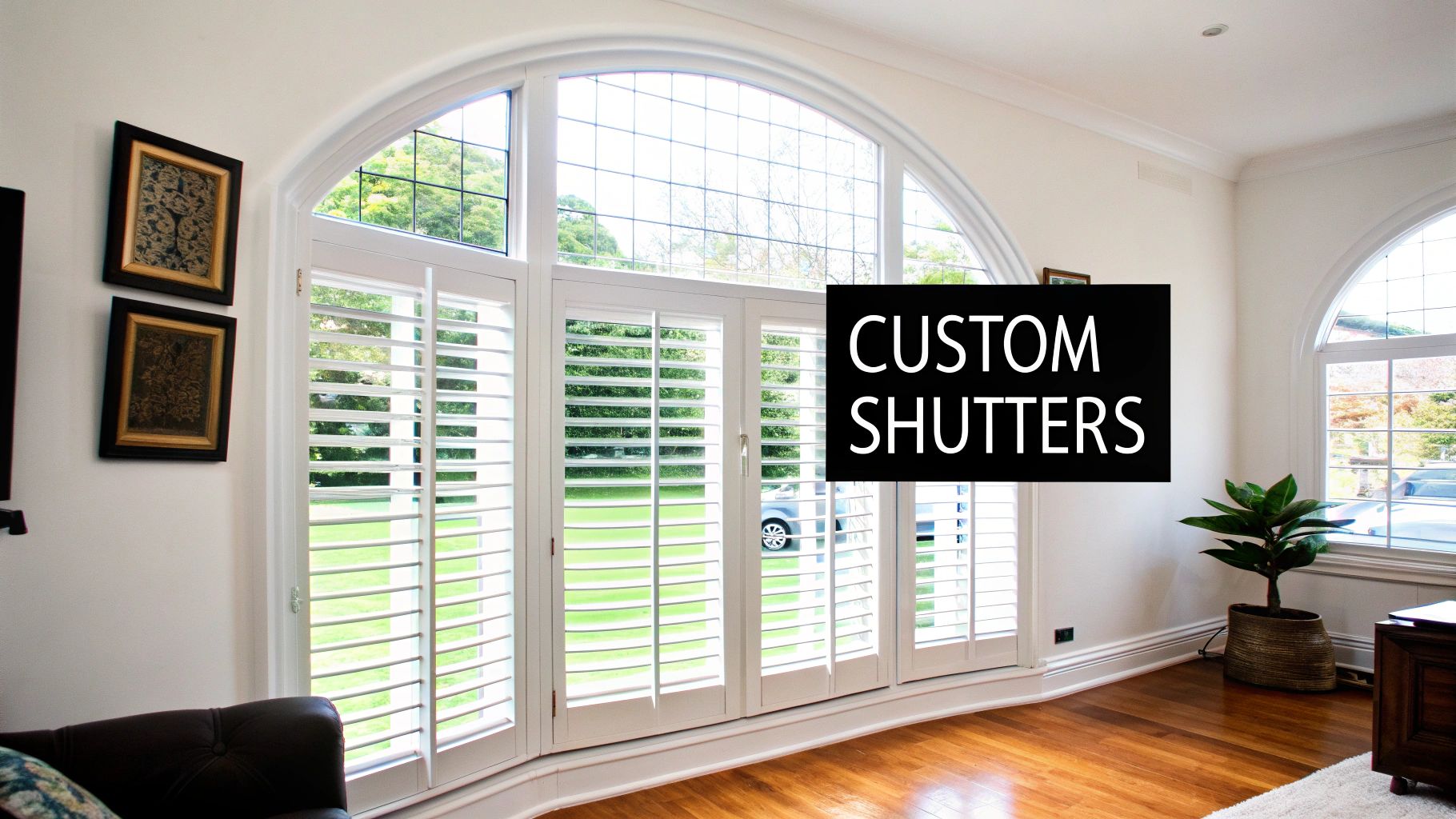A beautiful living room with custom white plantation shutters installed on a large curved bay window, letting in soft, filtered light.