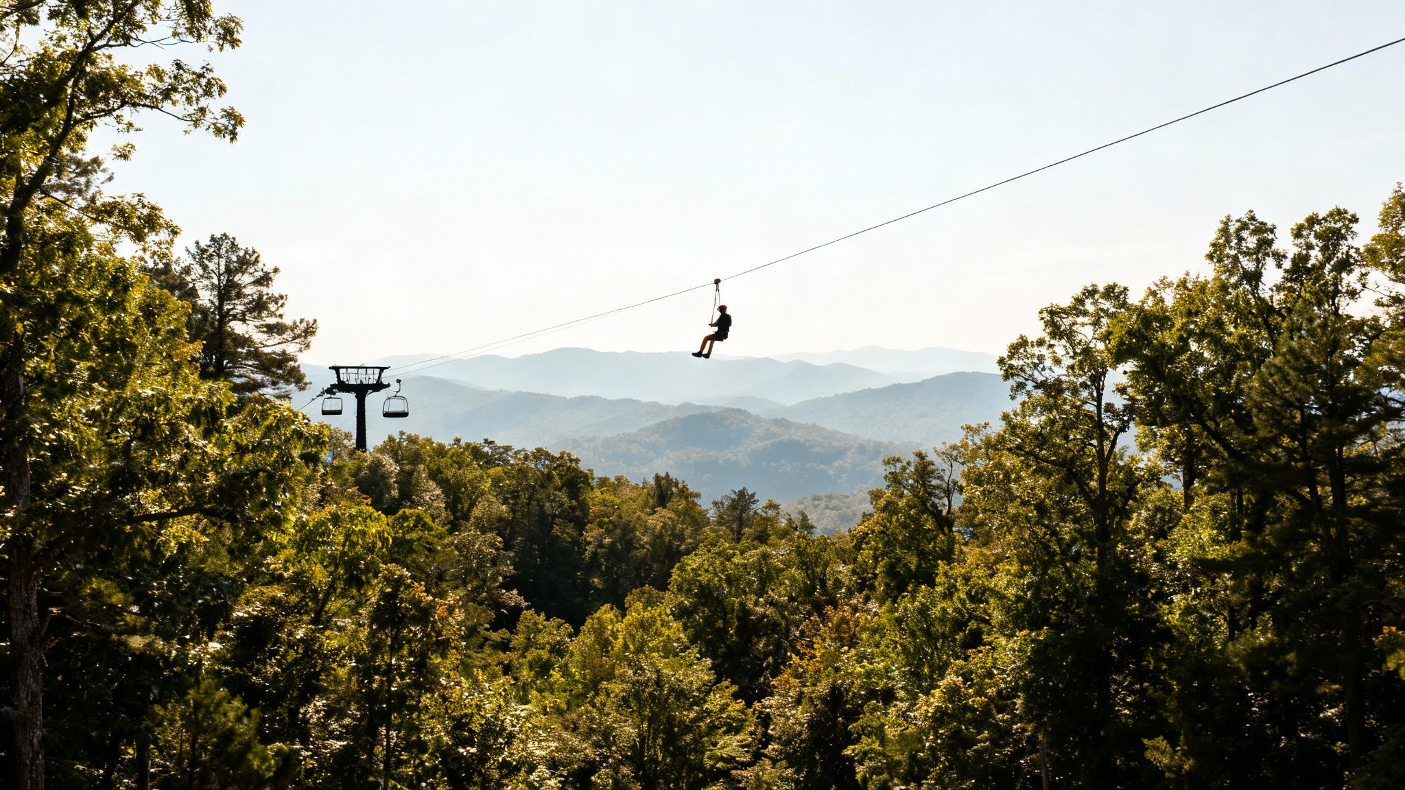 A person ziplining over lush green forests with misty mountains in the distance and a ski lift tower.