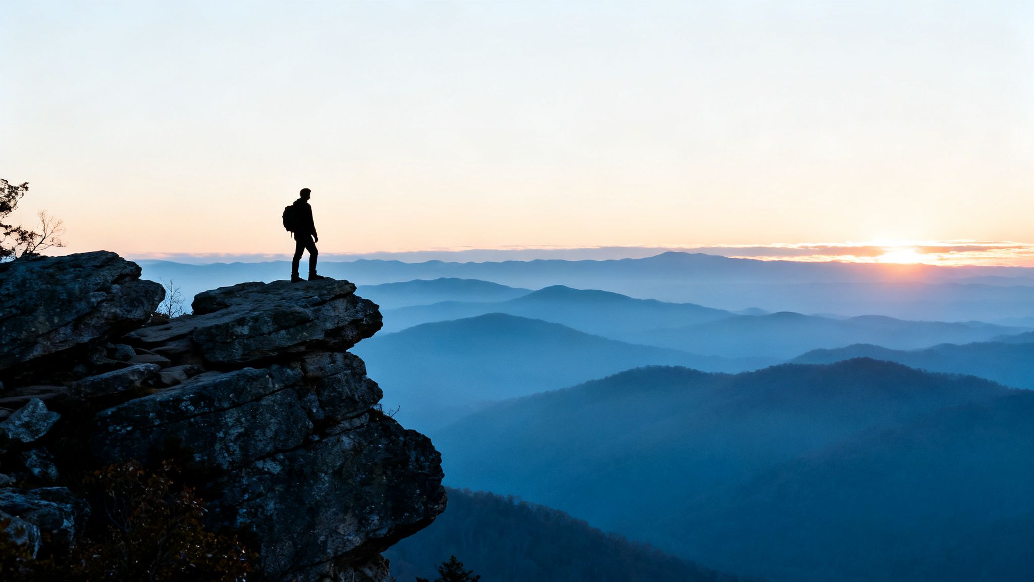 A solitary hiker stands on a mountain peak overlooking a vast, misty blue mountain range at sunset.