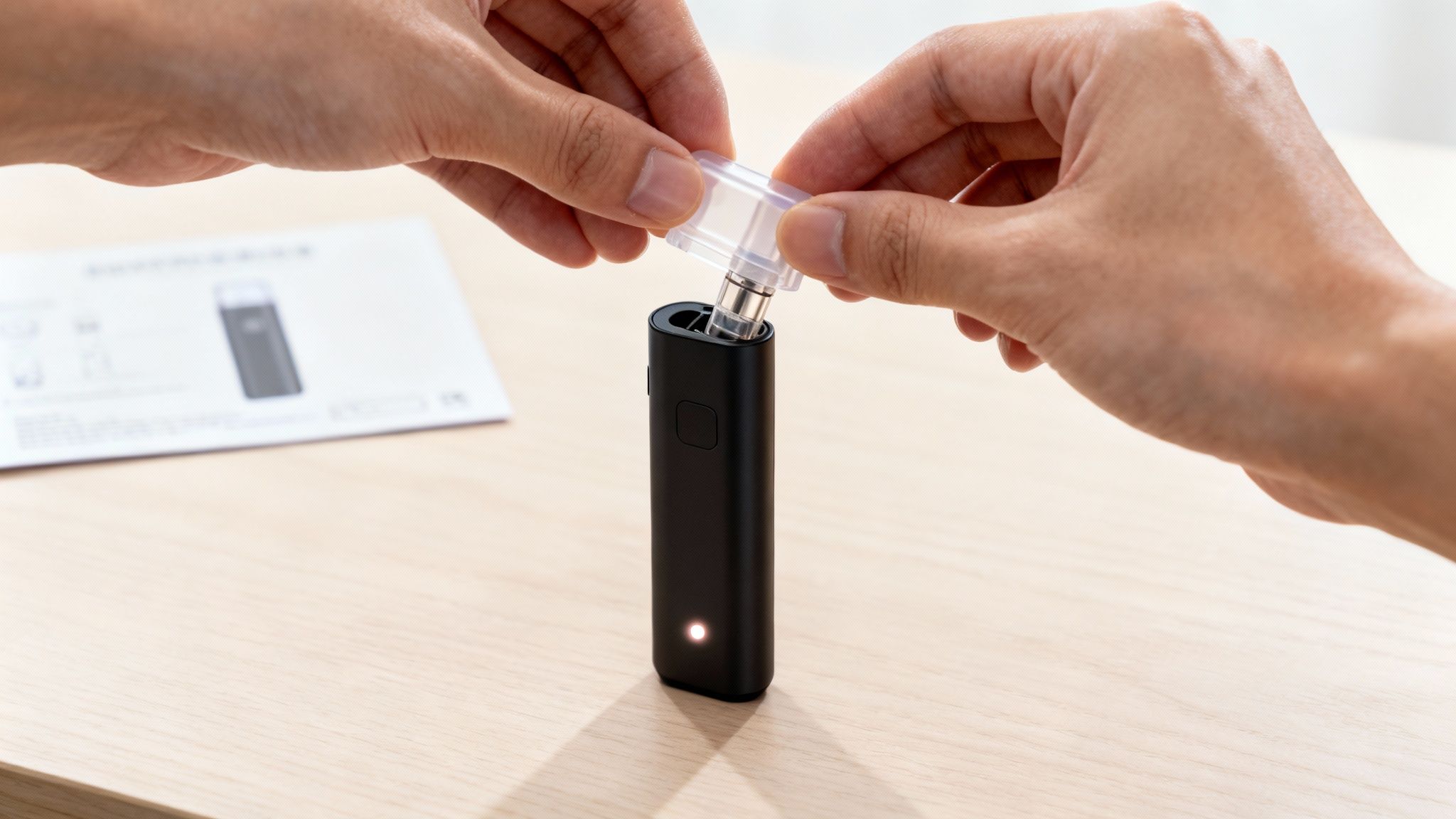 Hands insert a clear vape cartridge into a black electronic device with a glowing light on a wooden table.