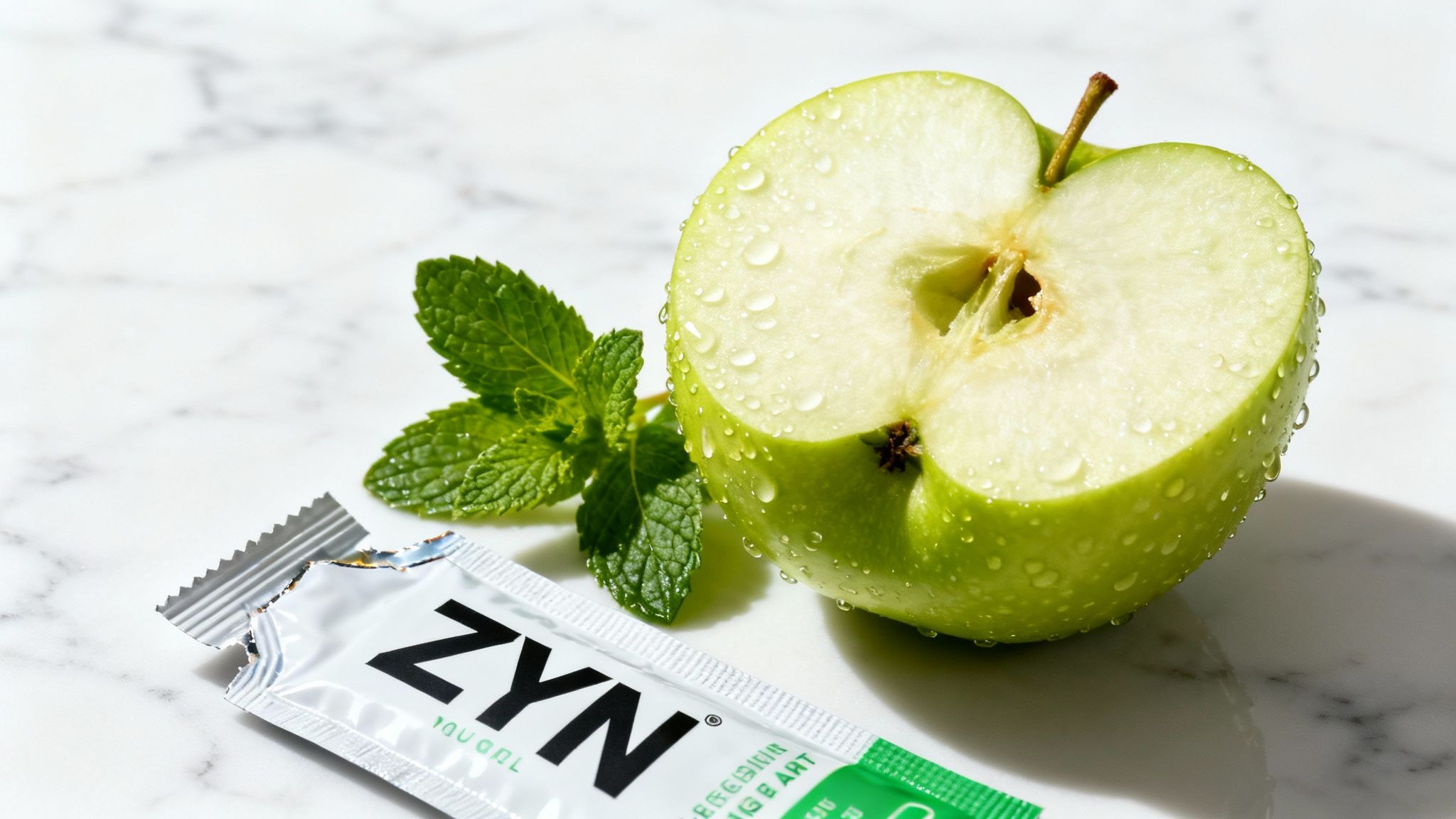 A fresh green apple half with water droplets, mint leaves, and a ZYN packet on marble.