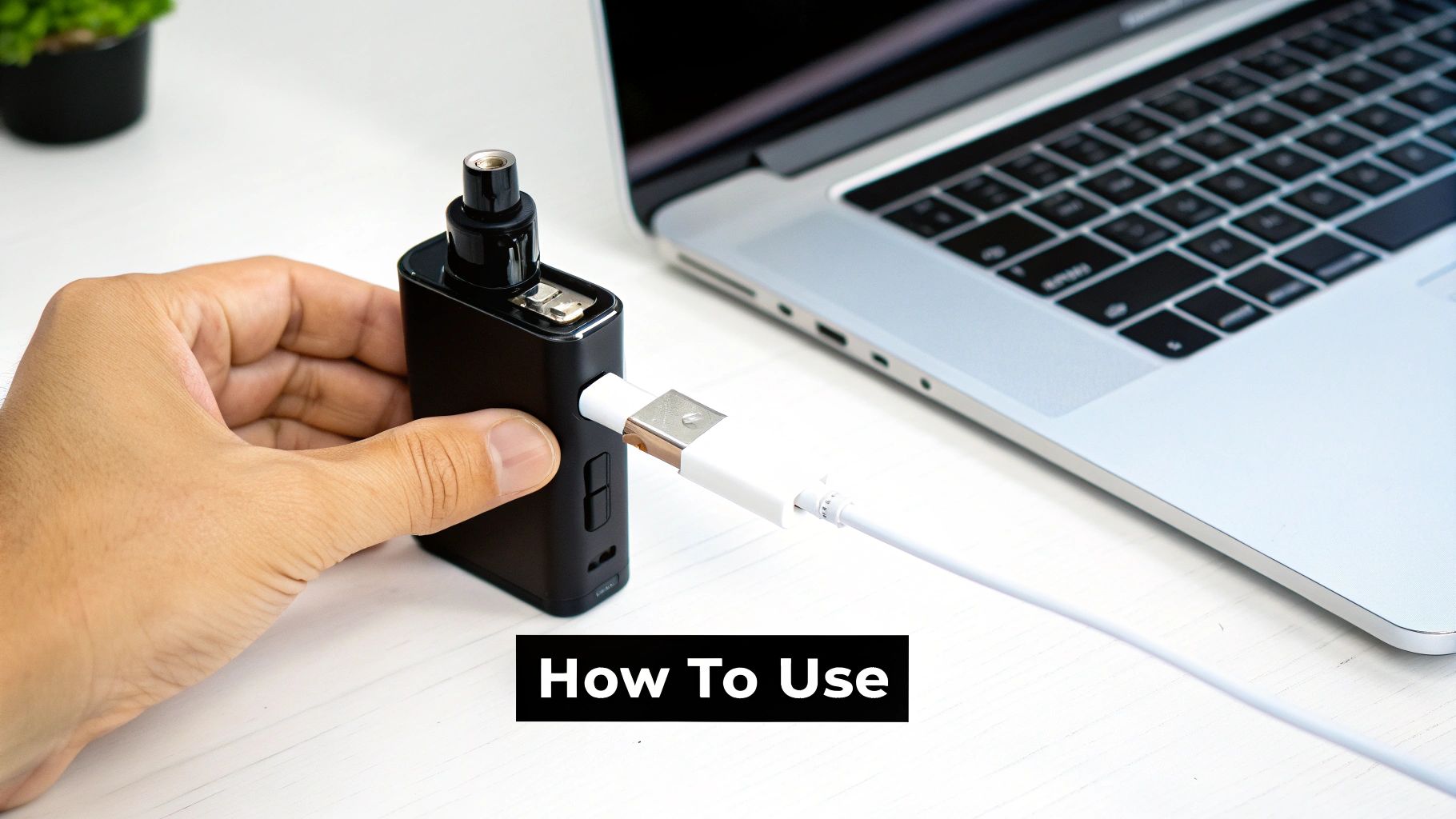 A person's hand holds a black vaping device, charging it via a white USB cable connected to a silver laptop.