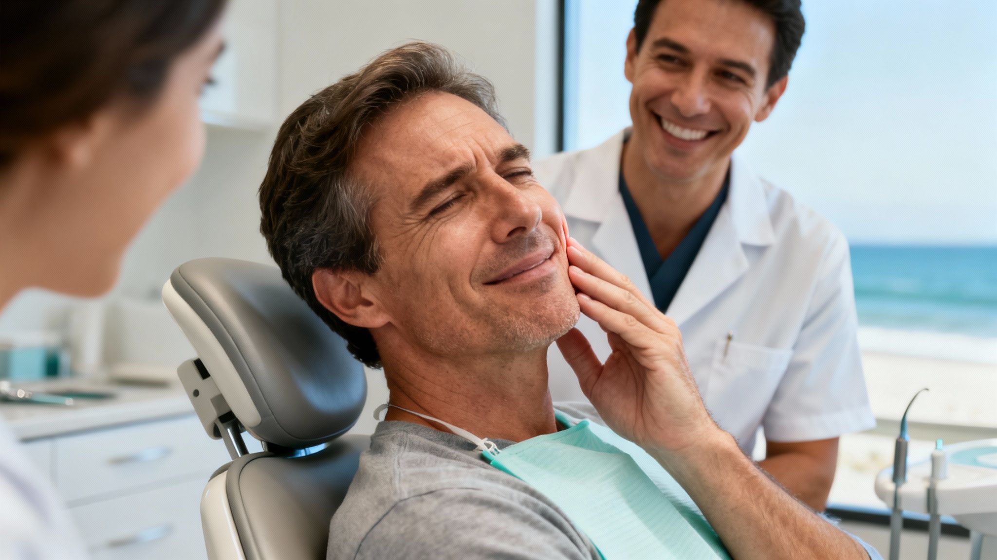 Man in dental chair touches his cheek while dentist smiles, with an ocean view.