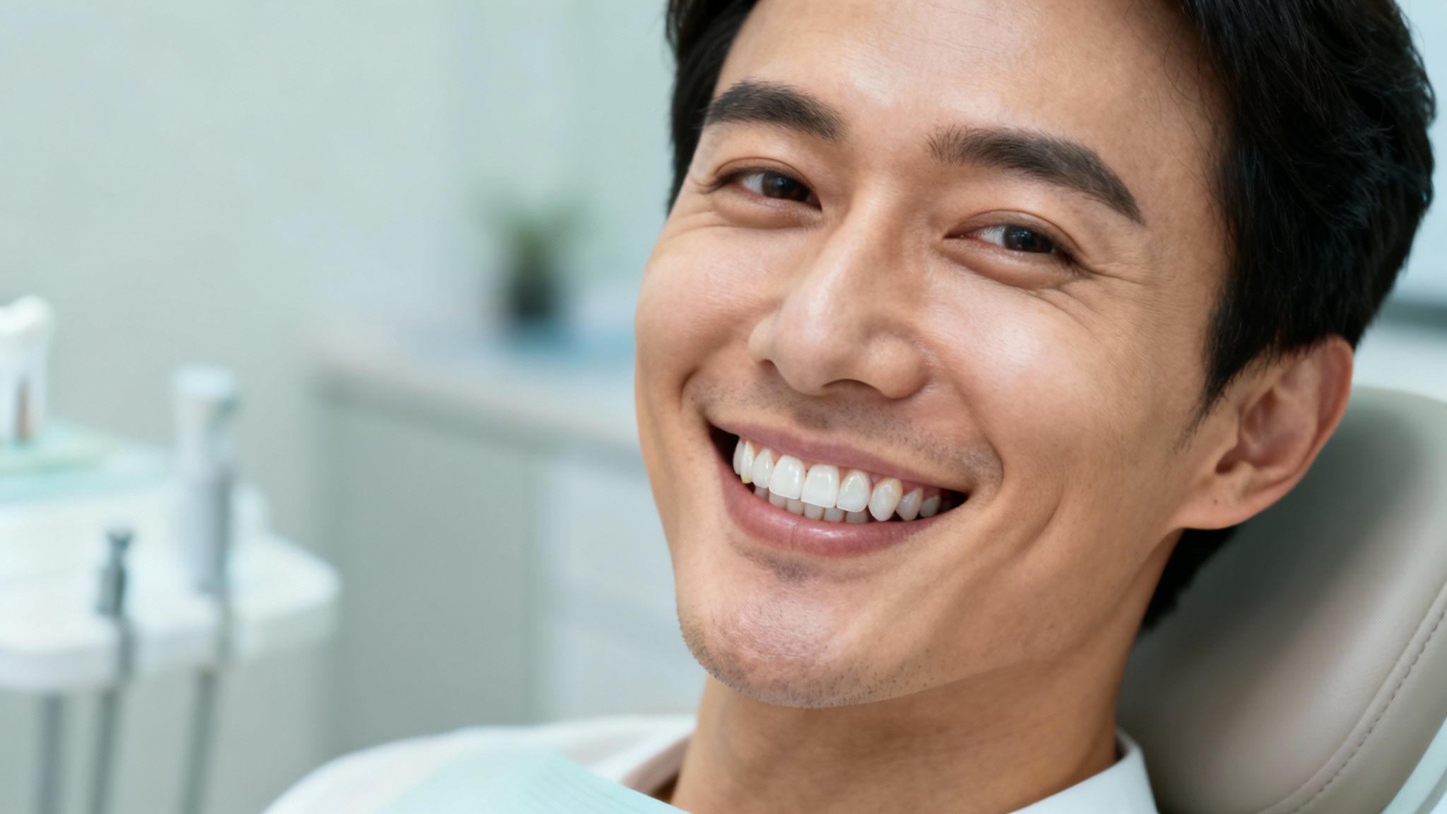 A smiling young Asian man with healthy, white teeth sits comfortably in a dentist's chair.