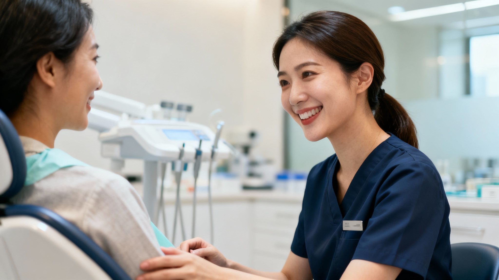A smiling Asian female dentist talks to a patient in a dental chair in a bright clinic.