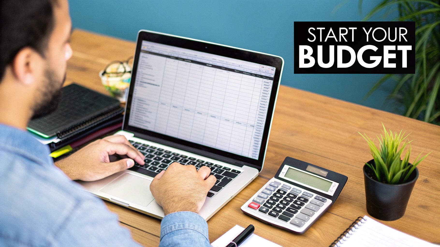 A person is typing on a laptop with a budget spreadsheet, calculator, and plant on a wooden desk.