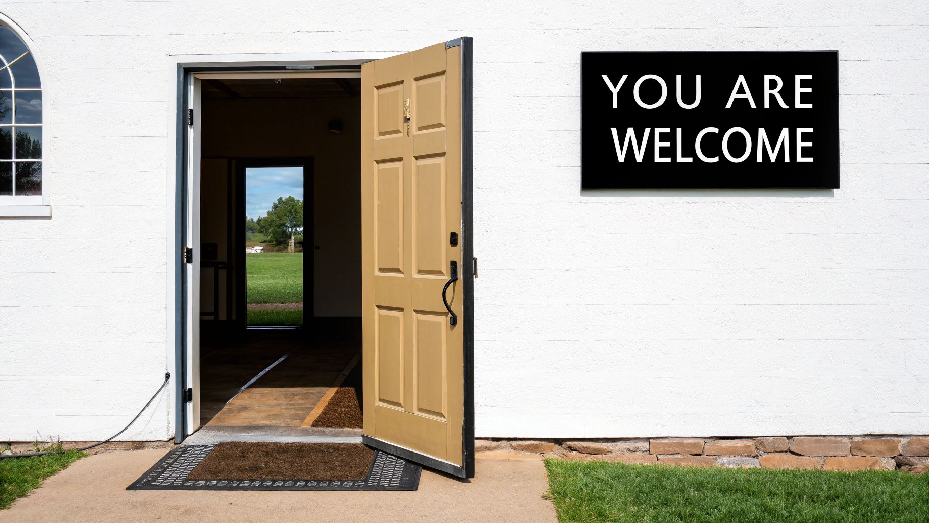 A white building with an open wooden door revealing a green field, next to a 'YOU ARE WELCOME' sign.