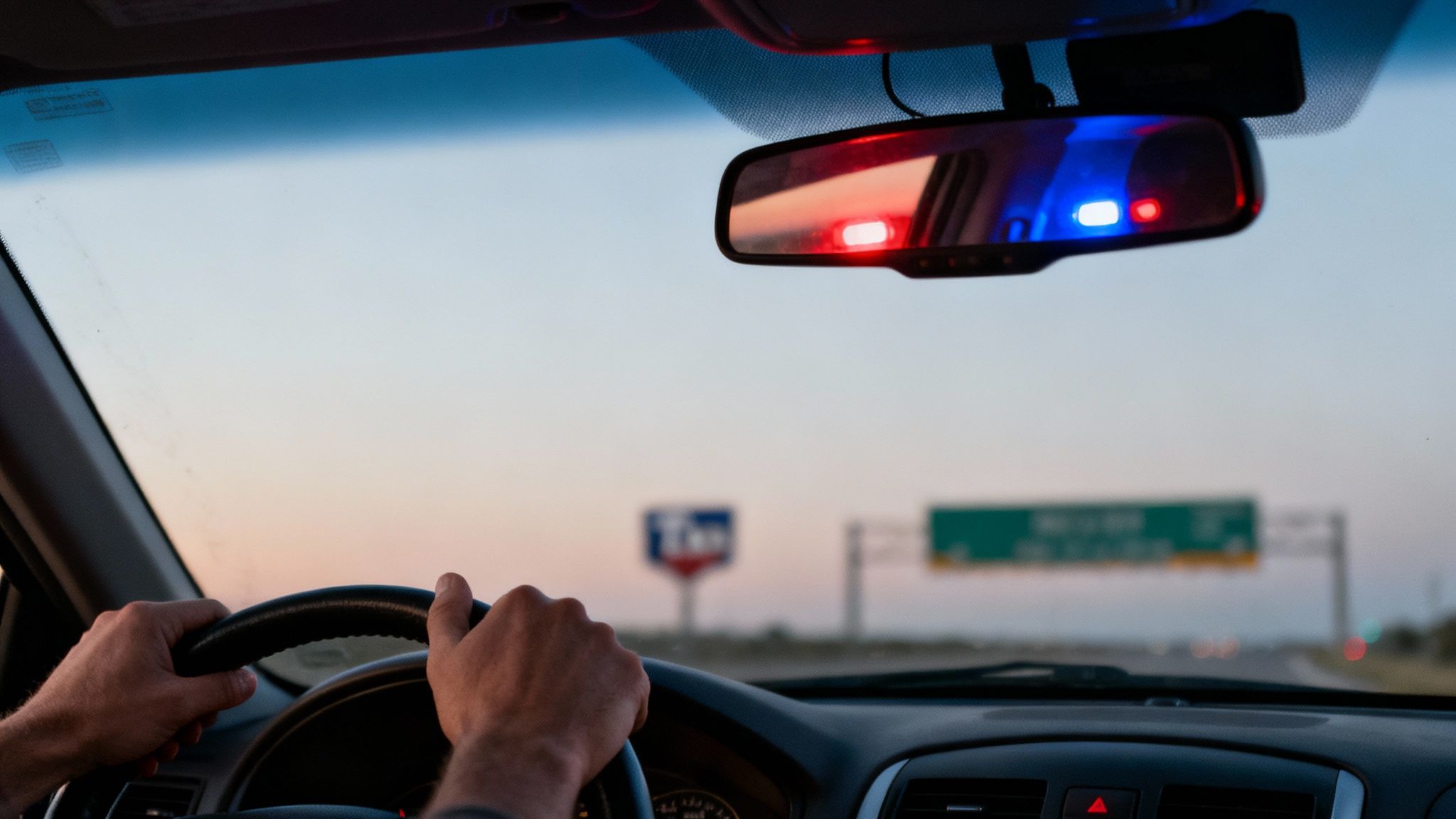 A driver's perspective from inside a car, with hands on the steering wheel and police lights reflected in the rearview mirror.