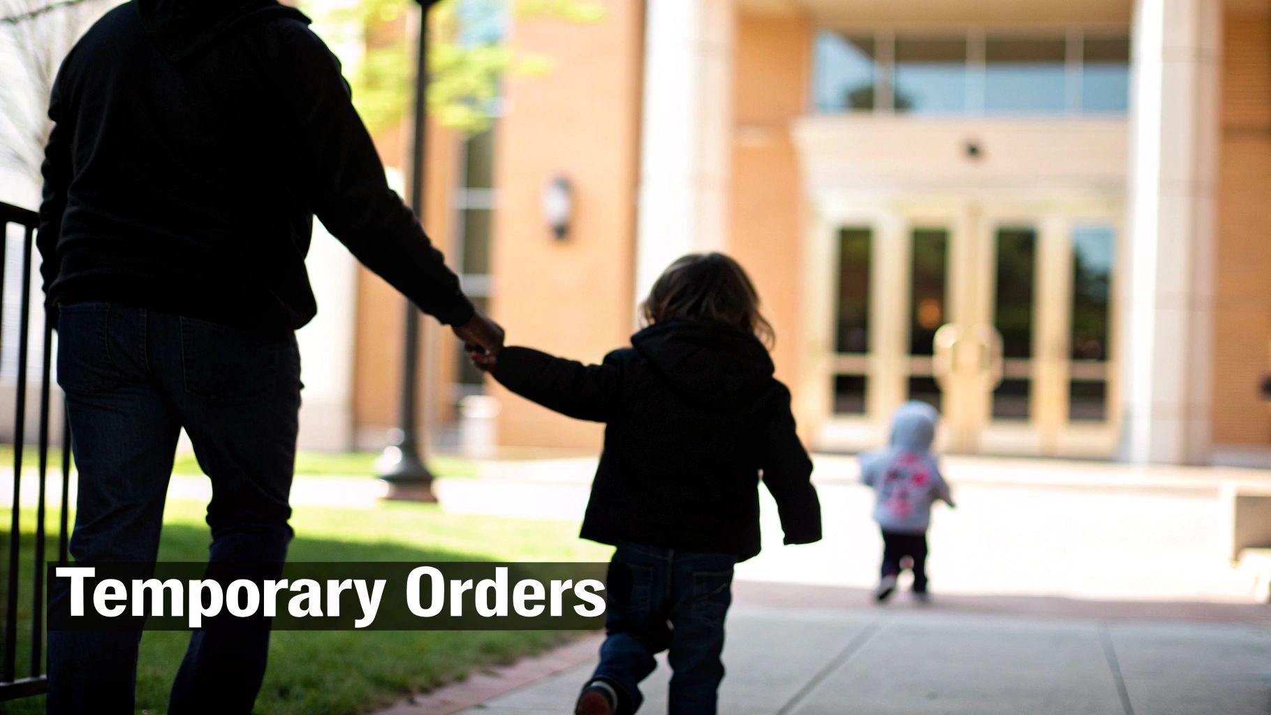 An adult holds a child's hand, walking away from the camera towards a blurred building, with another child ahead.