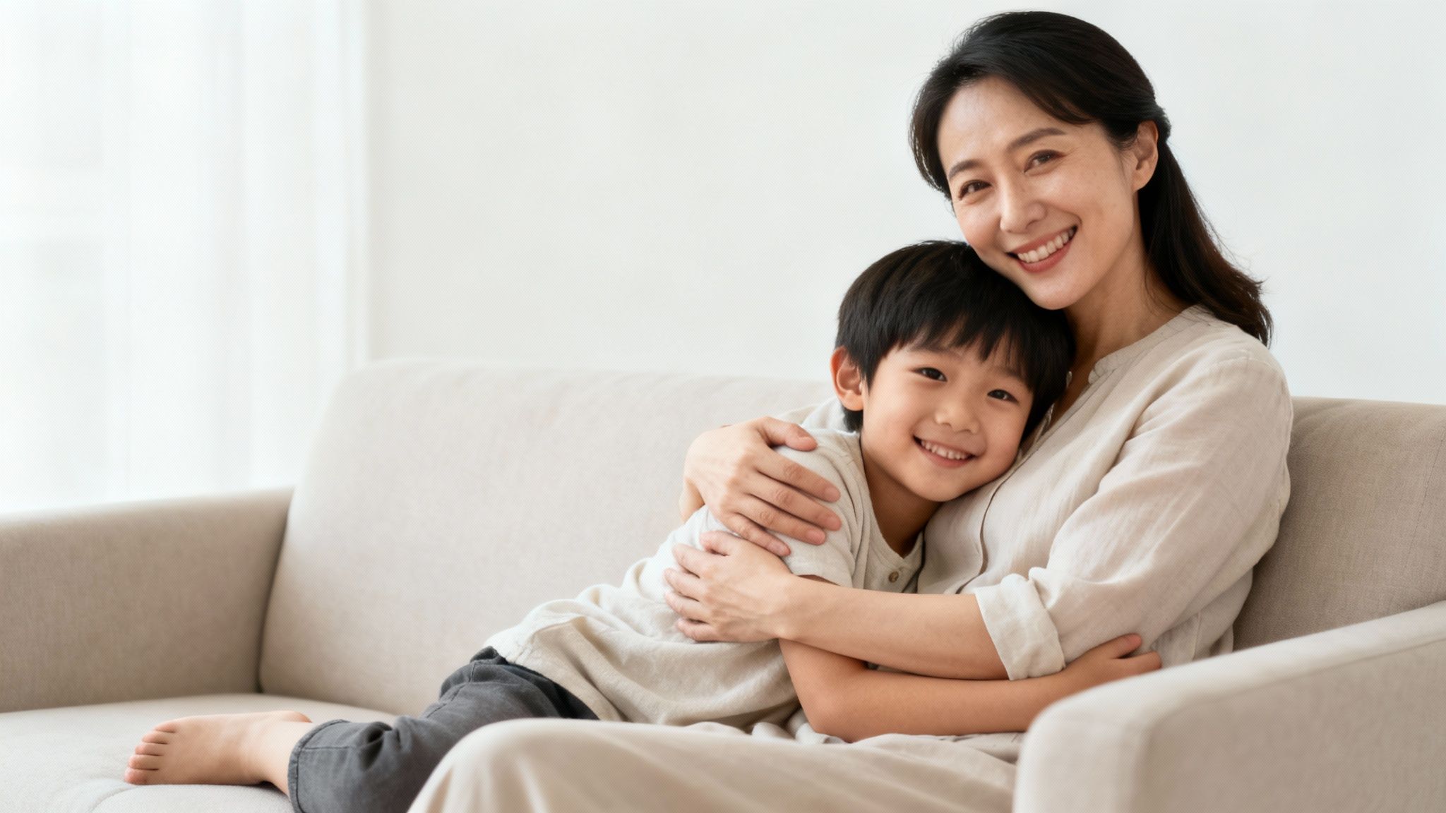 A loving Asian mother and her son embracing on a sofa, both smiling happily.