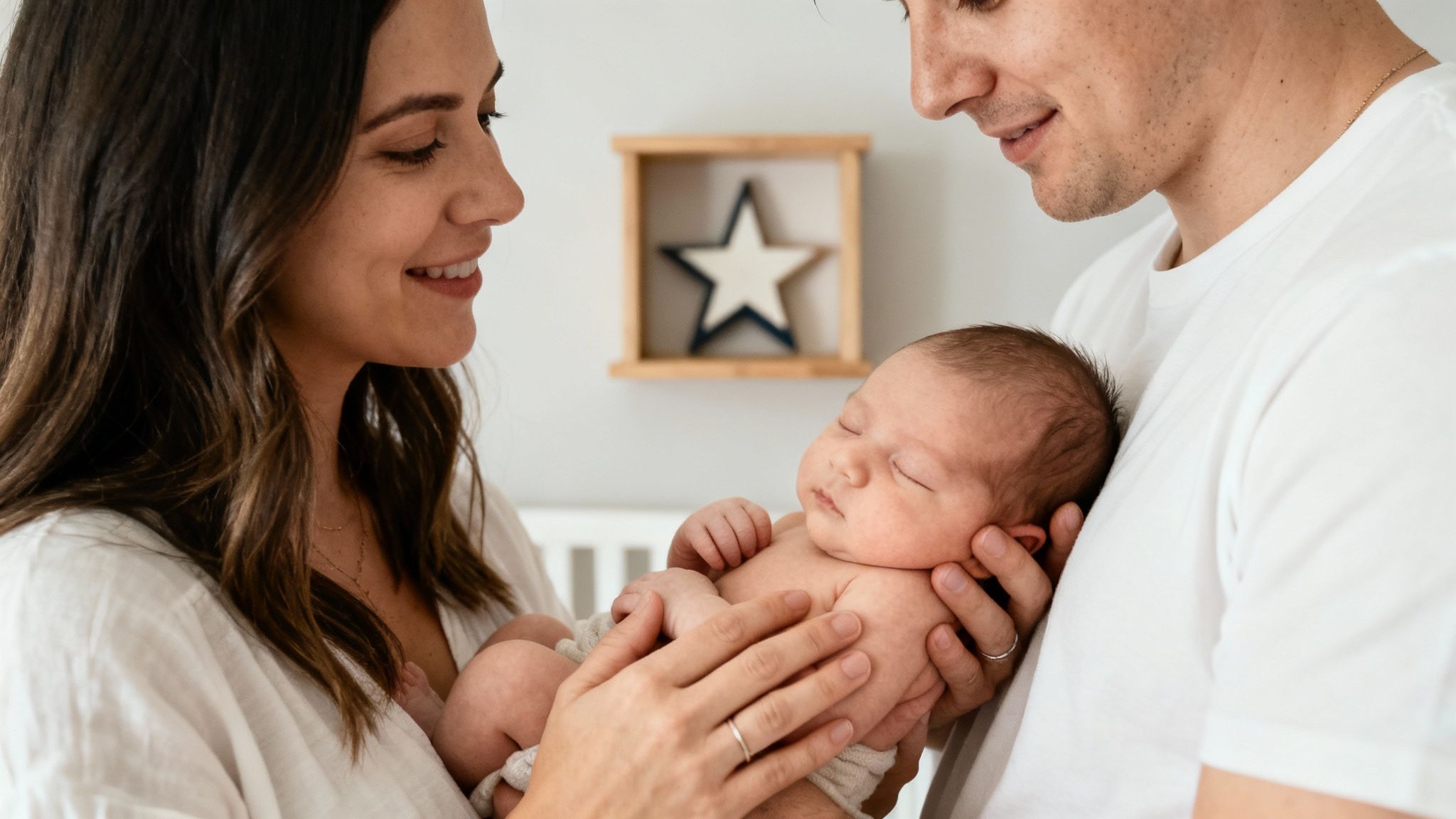 Smiling parents hold their sleeping infant in a warm embrace, looking at the baby lovingly.