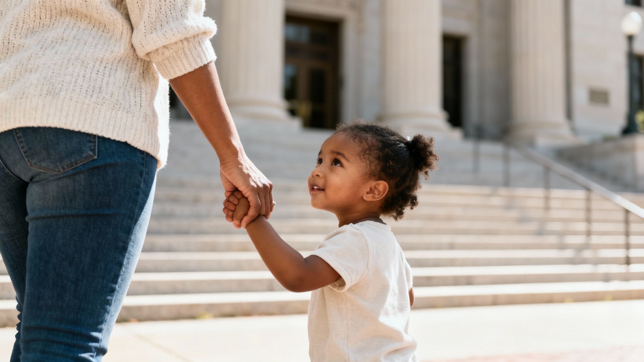 A young child gazes up happily, holding a parent's hand in front of a grand building.