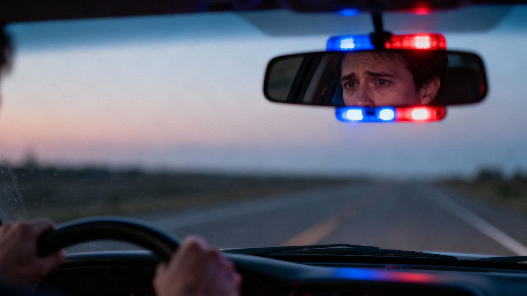 Worried driver's face reflected in rearview mirror with flashing police lights behind.