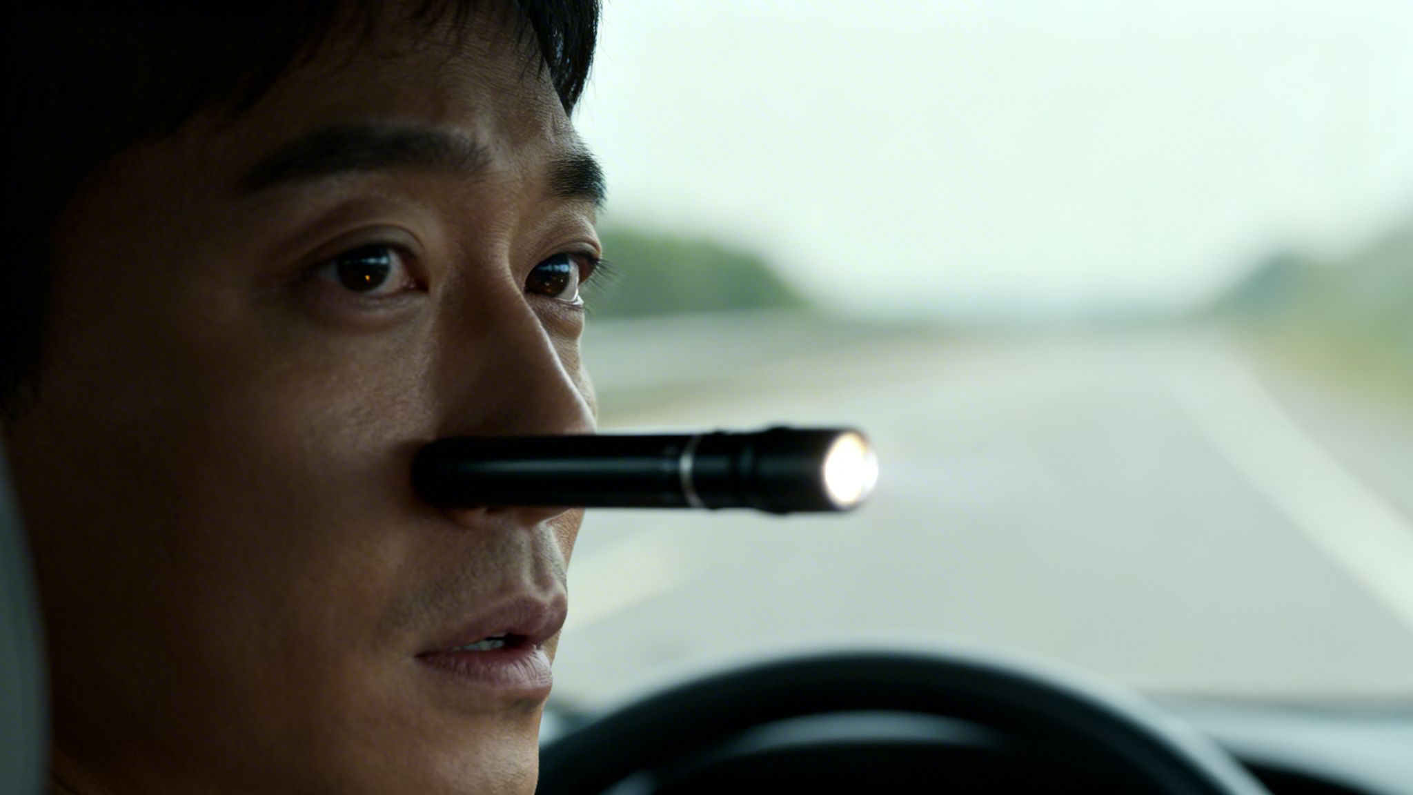 A man driving a car with a lit flashlight held under his nose, looking focused.