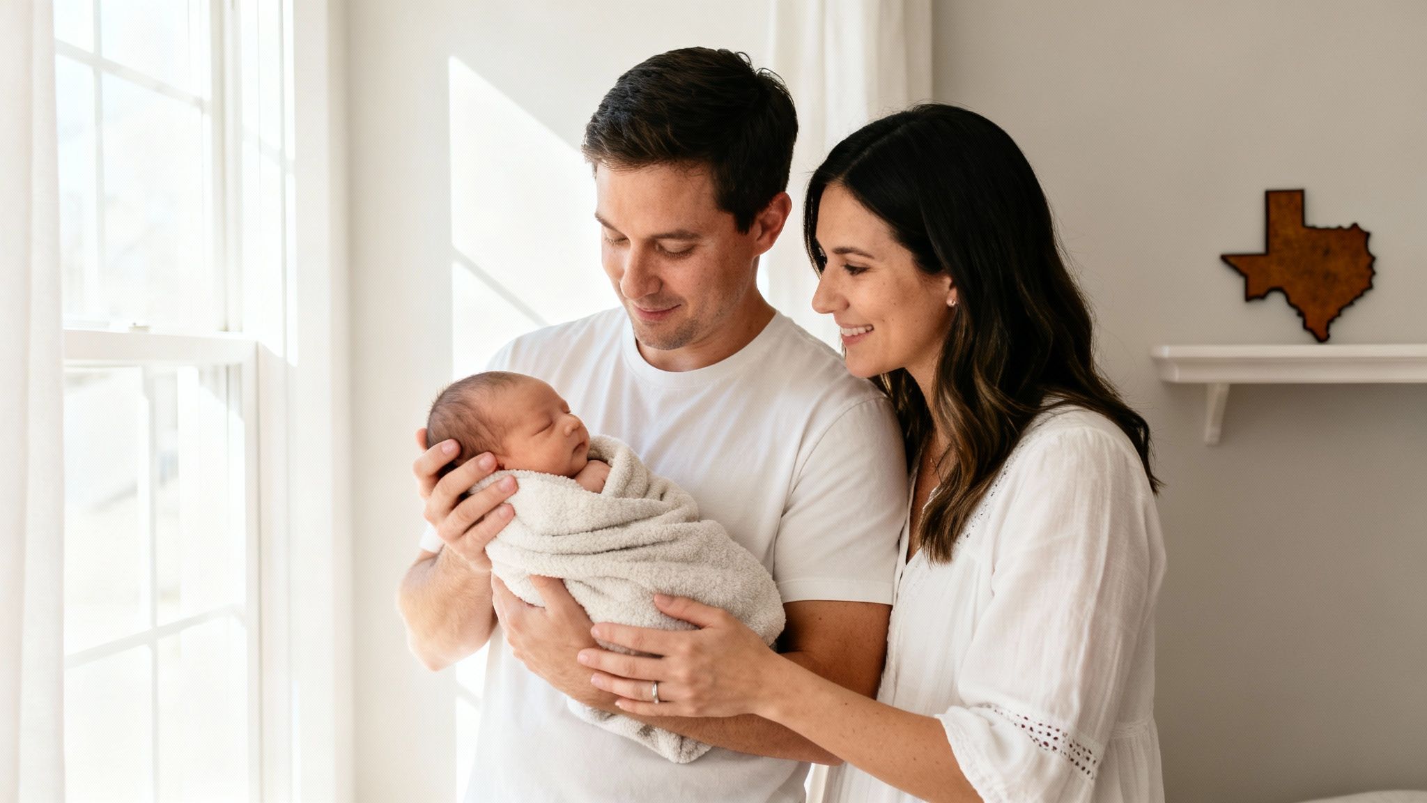 Smiling parents hold their swaddled newborn baby by a sunny window with a Texas decoration.