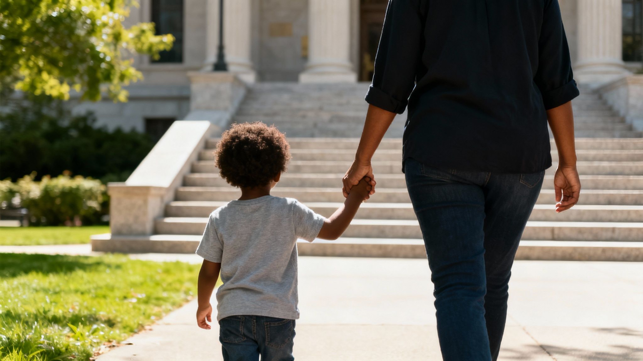 A child holding an adult's hand walks towards a grand building with steps on a sunny day.
