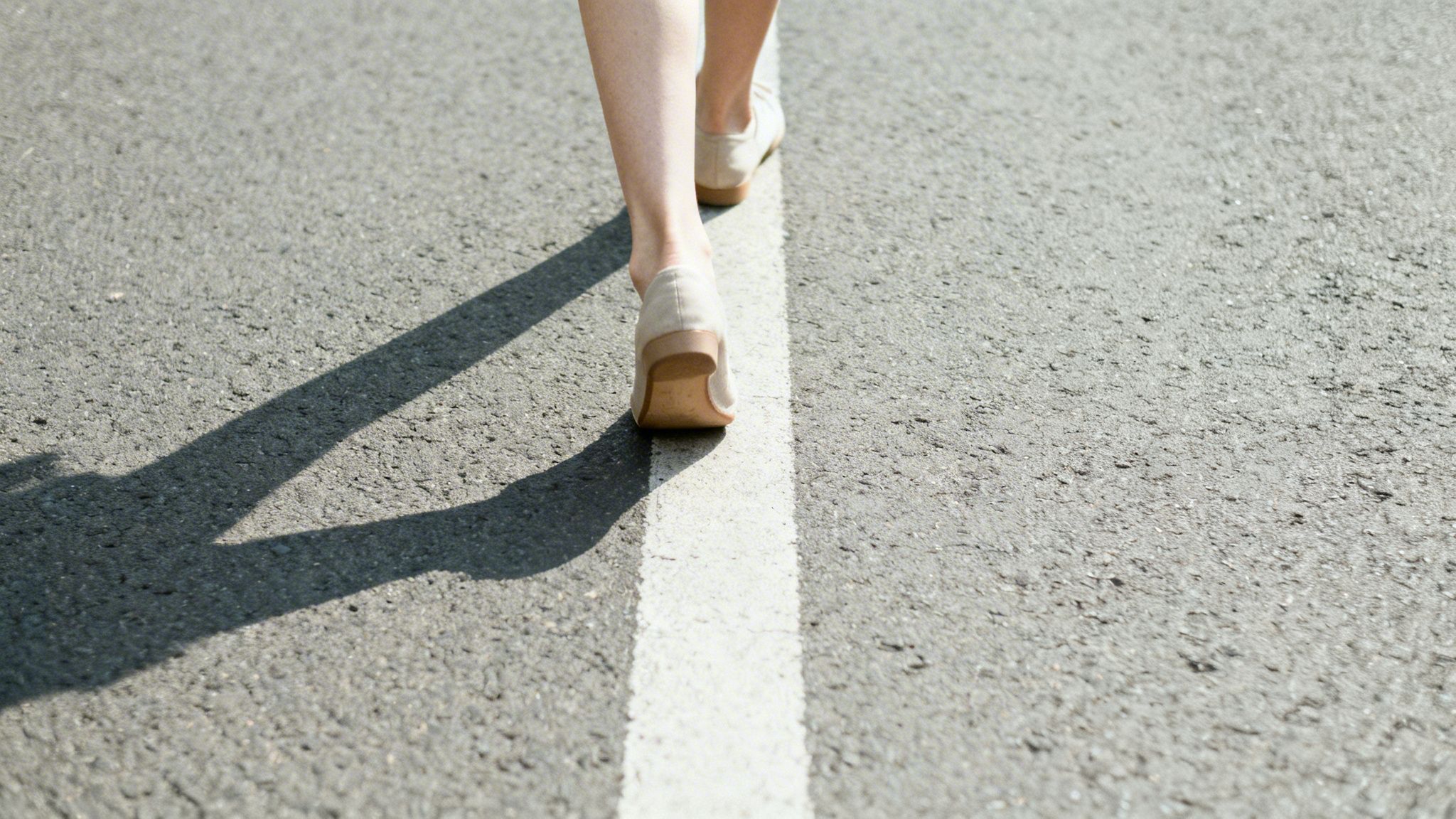 A person in light shoes walks on an asphalt road with a prominent white line and shadows.