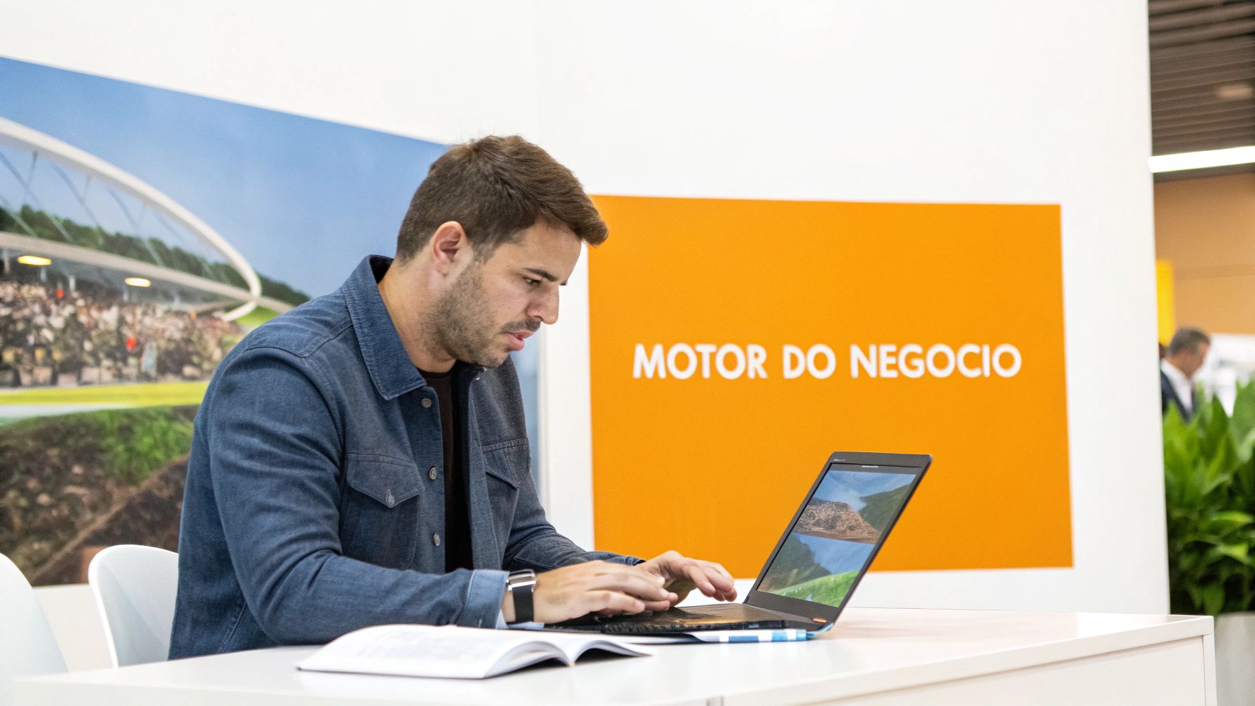 Homem focado em camisa jeans, usando laptop e lendo um livro em um ambiente profissional com um grande painel laranja ao fundo.