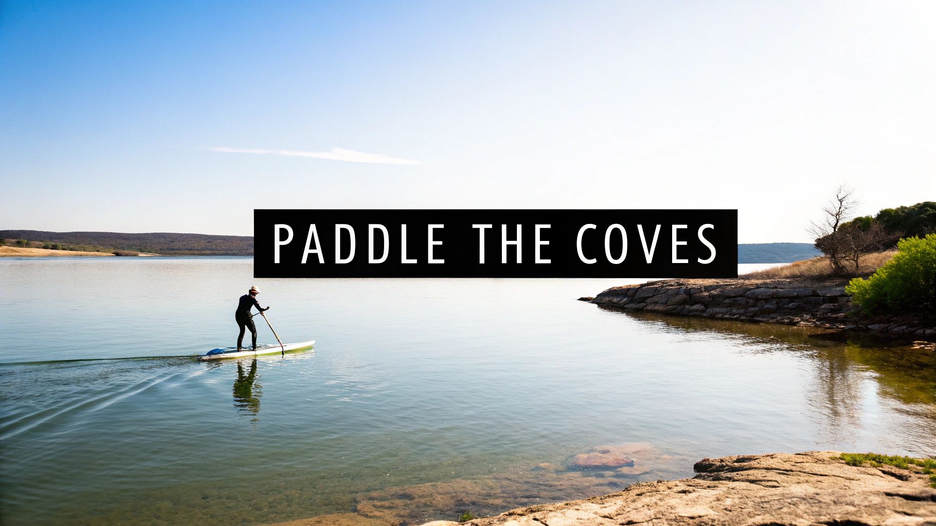 A person paddleboarding on a calm lake under a clear blue sky, with a rocky shore and distant hills.