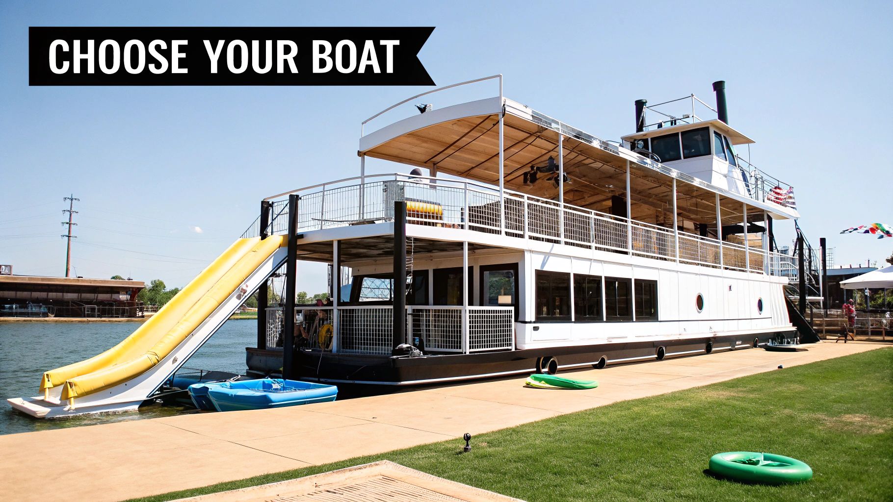 A large white party boat with a yellow water slide docked at a lake on a sunny day.