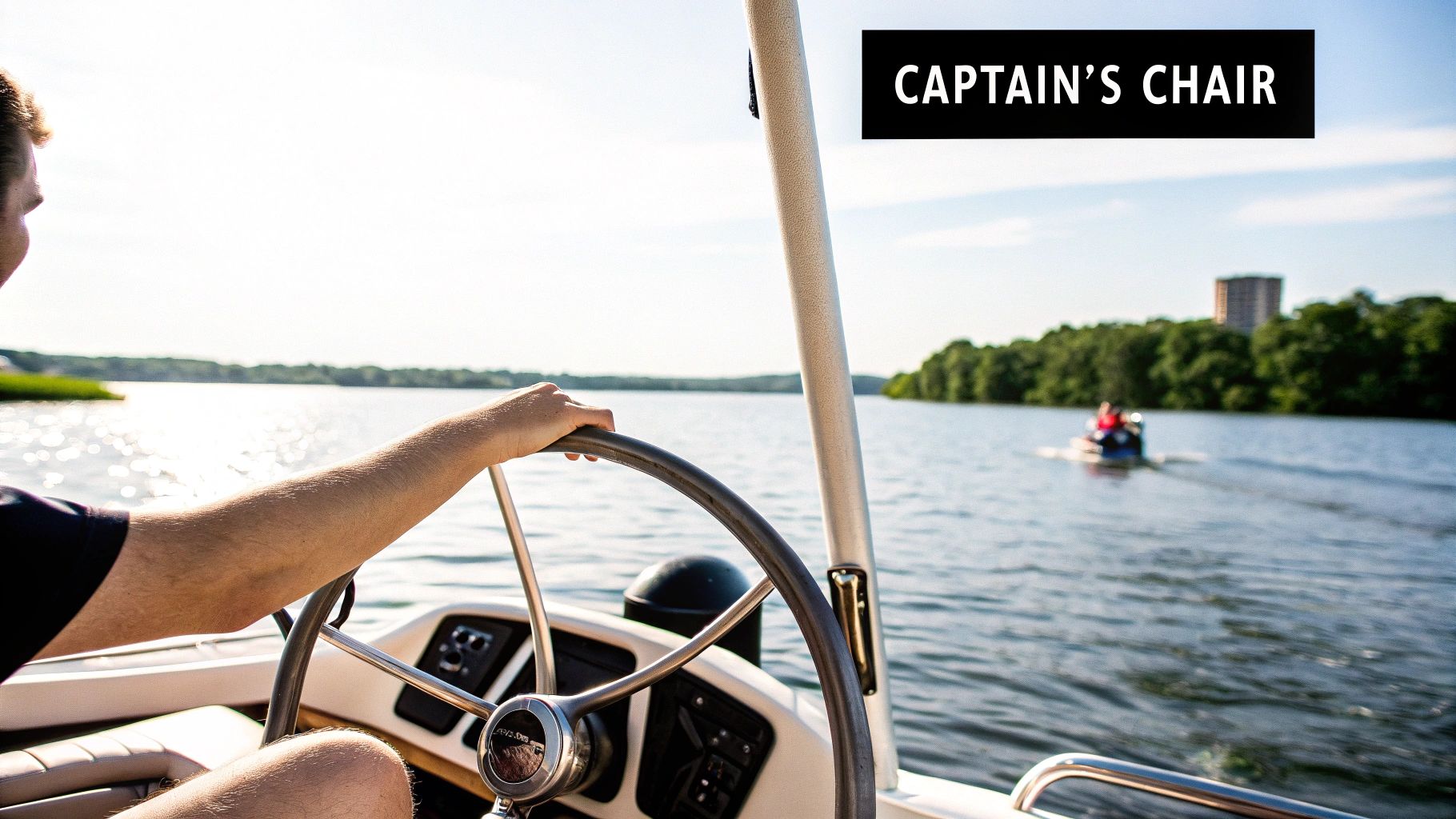 A person's arm on a boat's steering wheel, looking out at a sunny lake with another boat and distant shore.