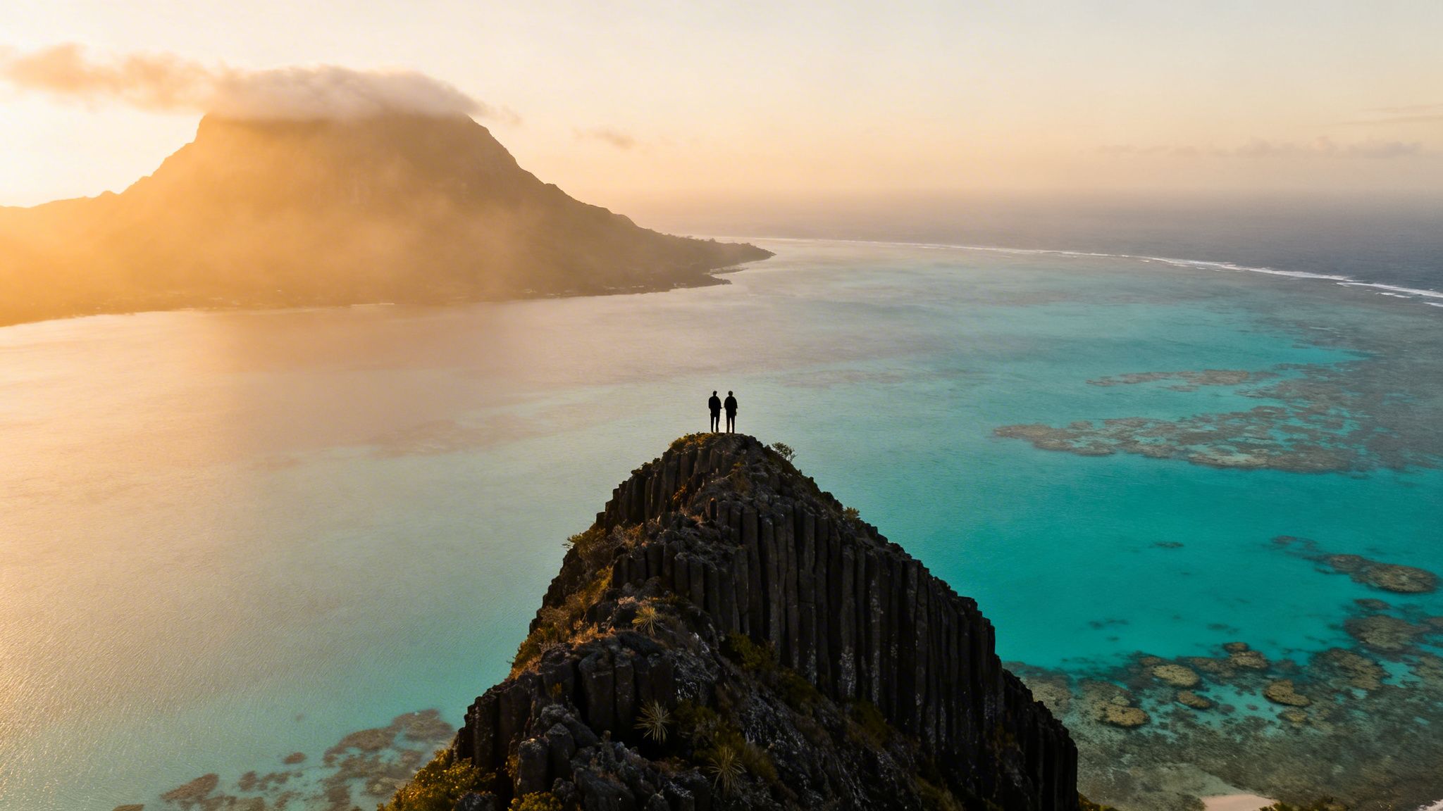 Two people stand on a rocky peak, admiring the turquoise lagoon and misty mountains at sunset in Mauritius.