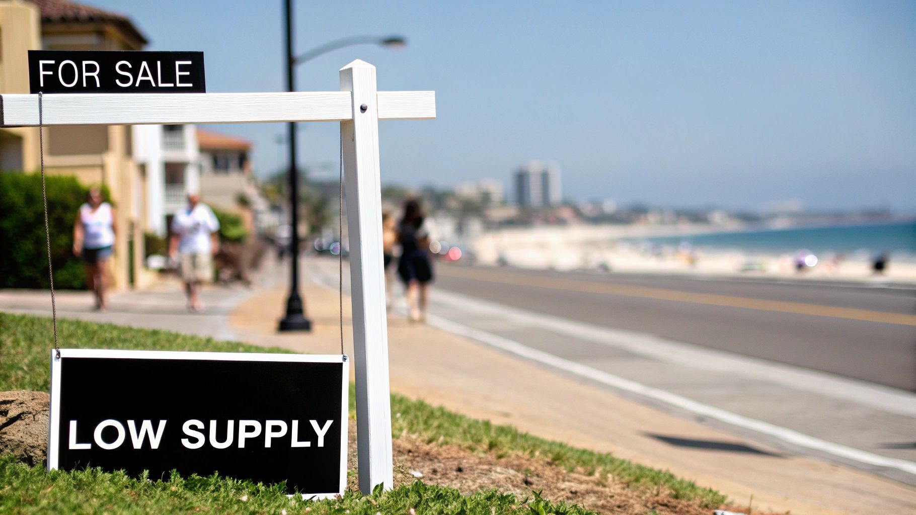 A real estate 'FOR SALE' sign highlights 'LOW SUPPLY' on a sunny day by a coastal road and beach.