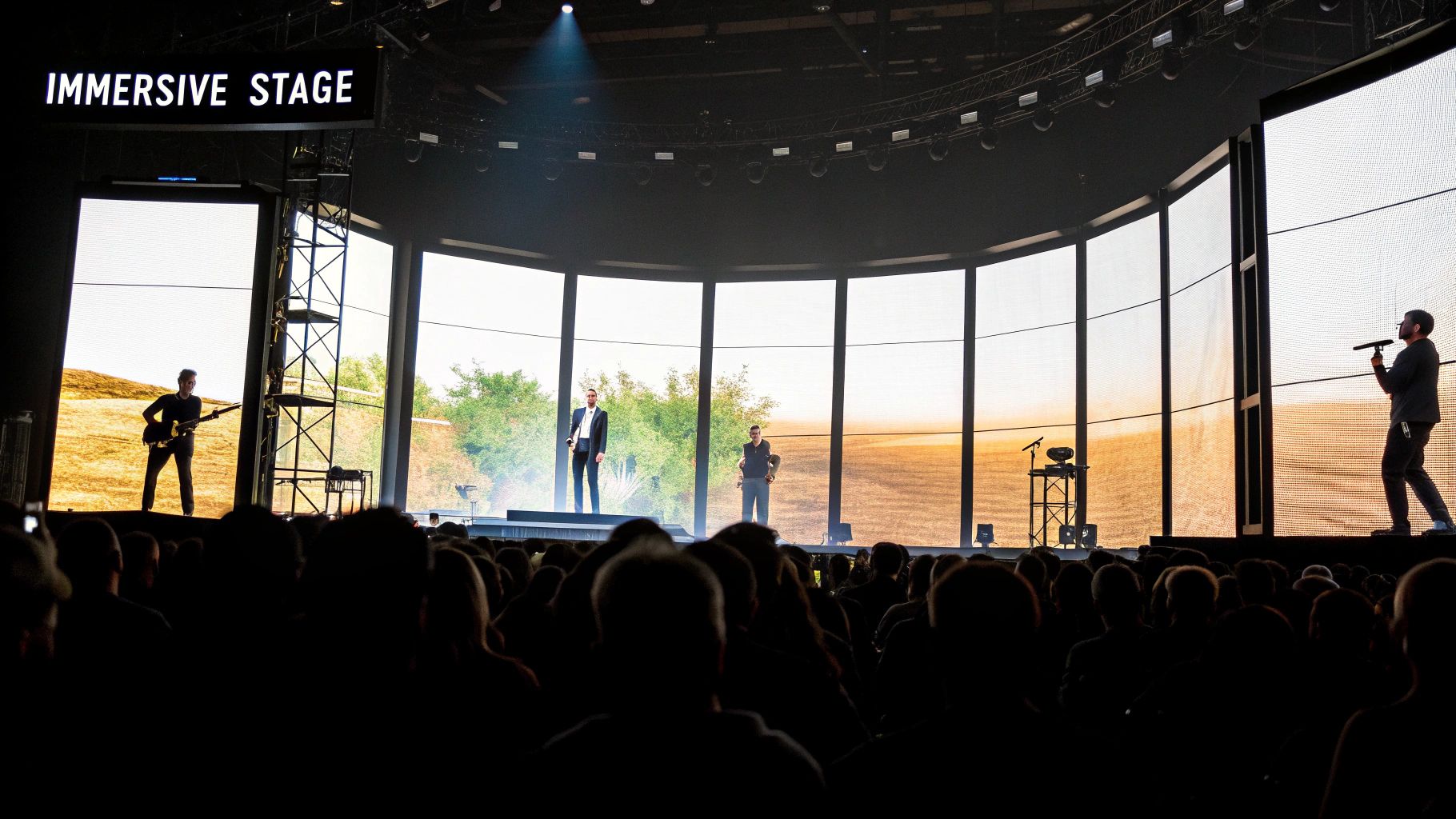 A band performs on an immersive stage with large LED screens displaying outdoor landscapes to a crowd.