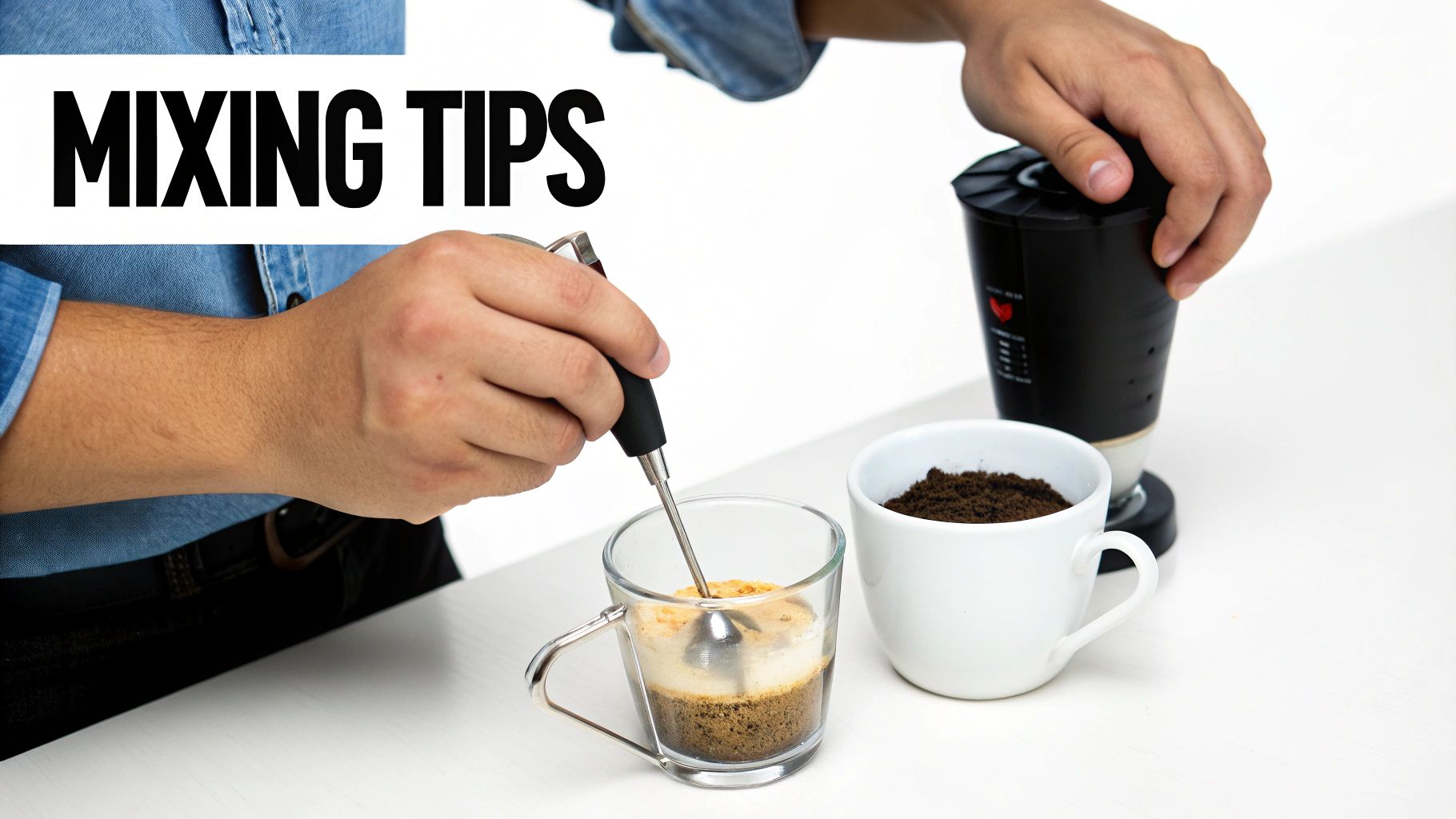Person mixing a coffee drink in a glass mug with a frother, alongside coffee grounds and a grinder.