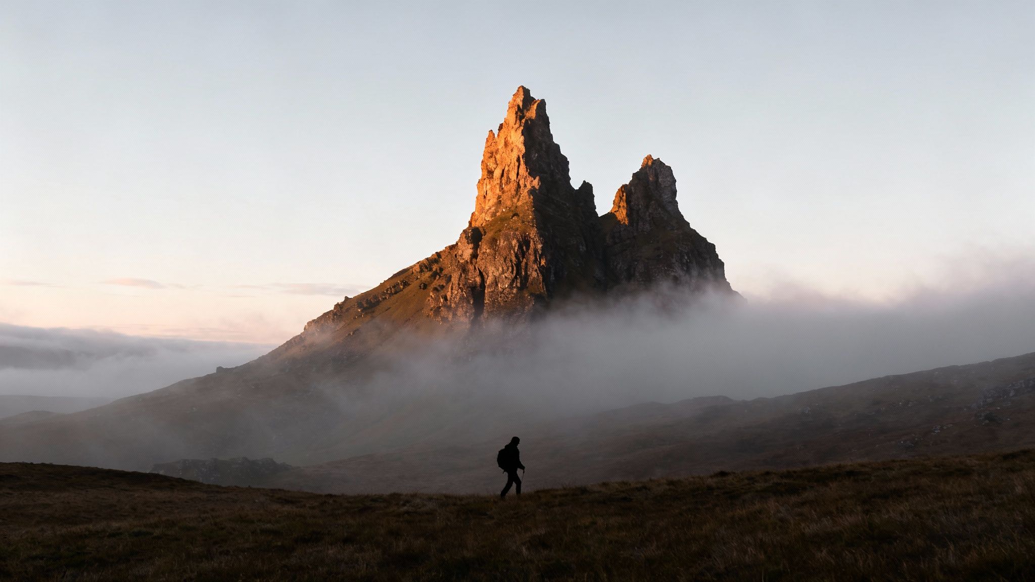 A lone hiker walks across a field towards majestic, mist-shrouded mountains bathed in golden light.