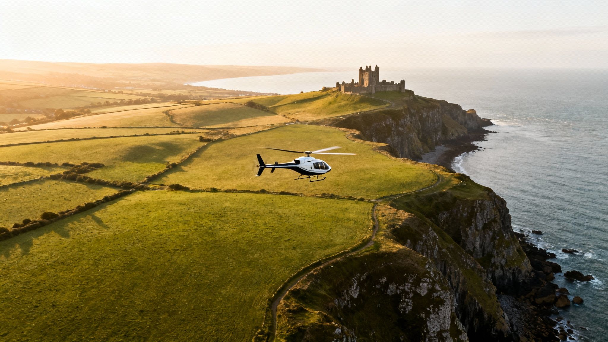 A white helicopter flies over lush green fields near a coastal castle at sunset.