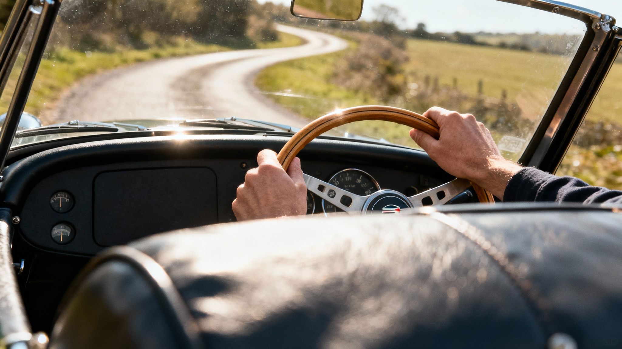 Close-up of hands on a wooden steering wheel of a classic car driving on a sunny winding country road.