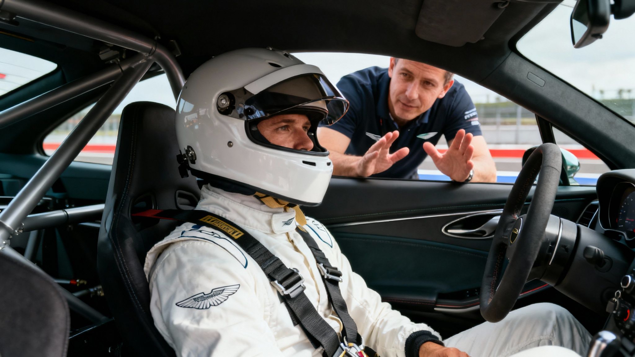 A race car driver in an Aston Martin, wearing a helmet and suit, listens to an instructor on the track.