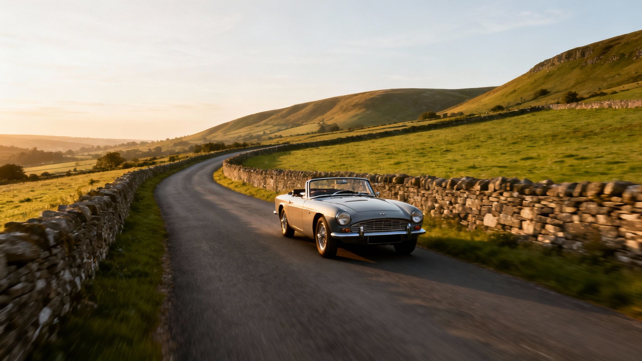 A classic grey convertible car drives on a winding rural road between green hills at sunset.
